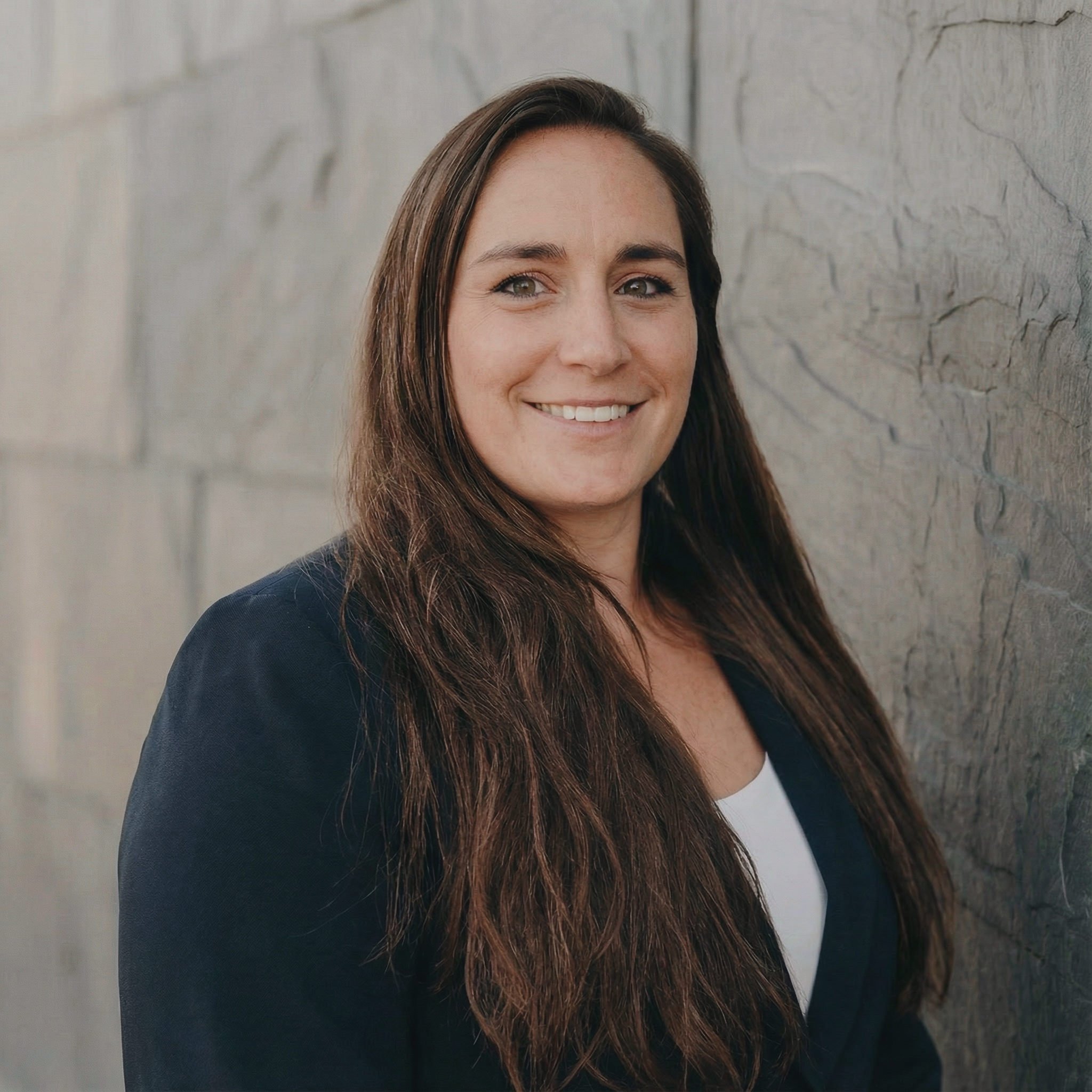 Woman with long brown hair smiling, wearing a dark blazer and white shirt, standing against a textured stone wall.