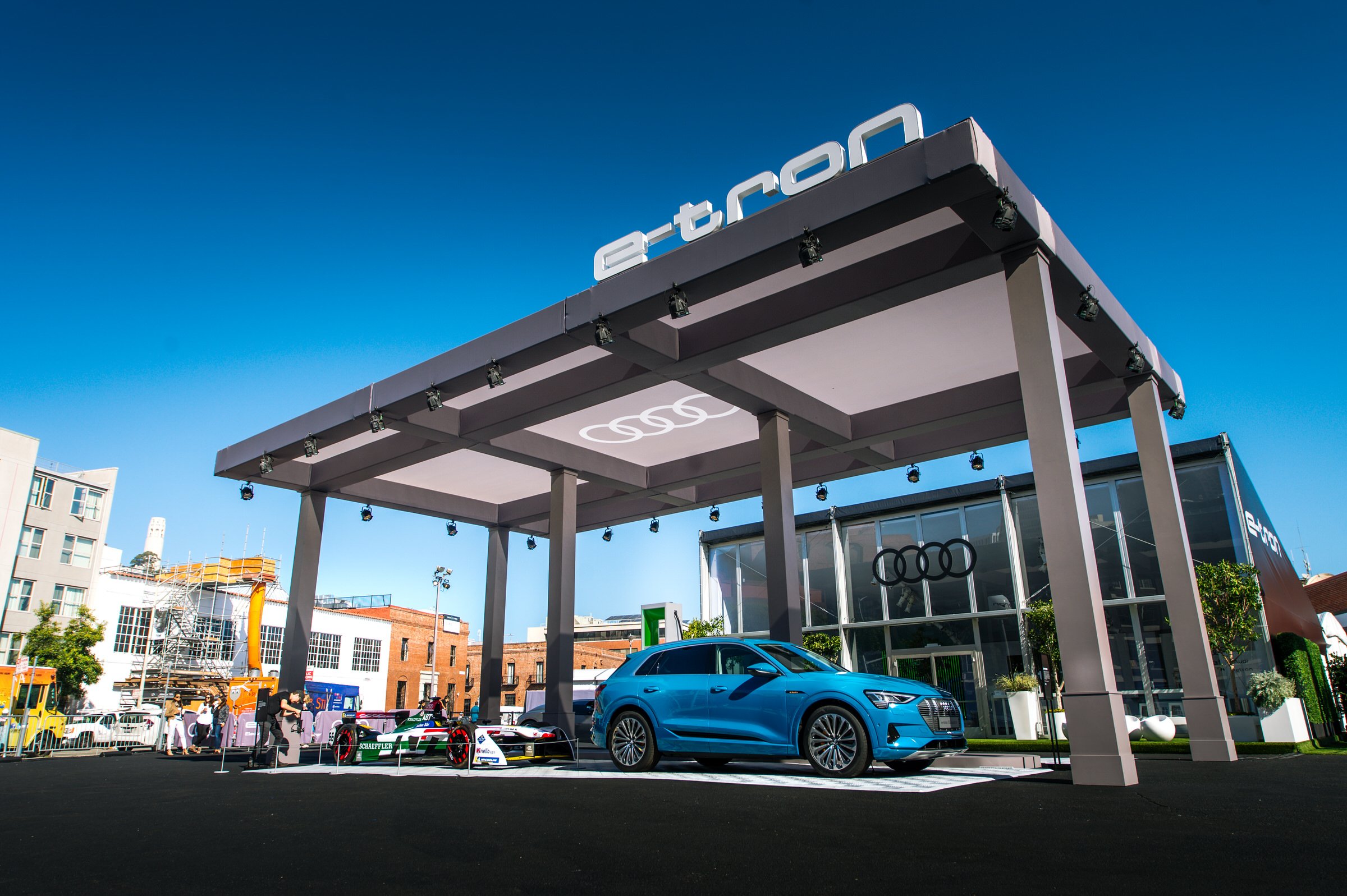 A car exhibition featuring an Audi vehicle and racing cars under a modern canopy with the Audi logo on a glass building, set against a clear blue sky.