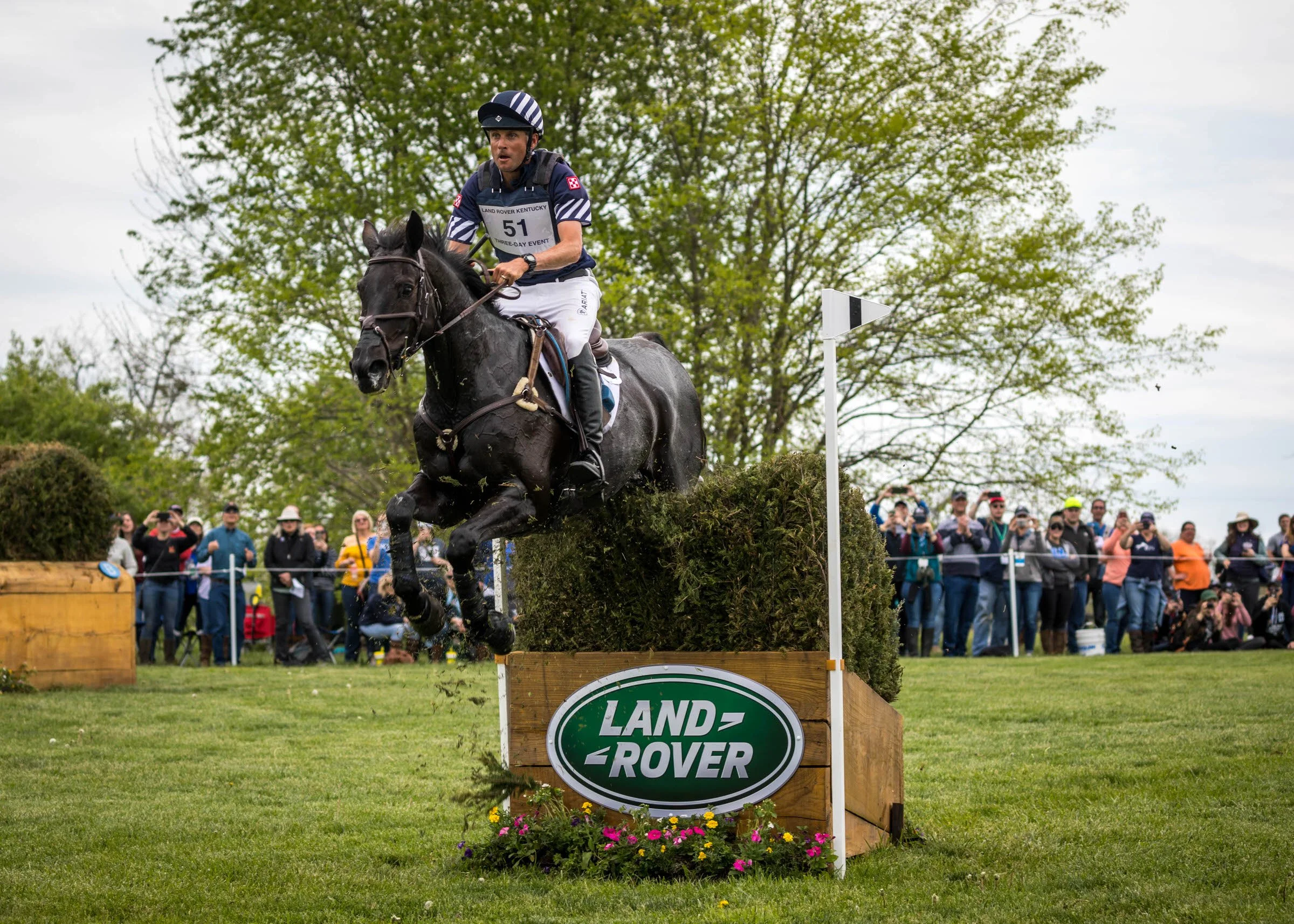 A person riding a horse jumps over an obstacle during an equestrian event, with spectators watching in the background.