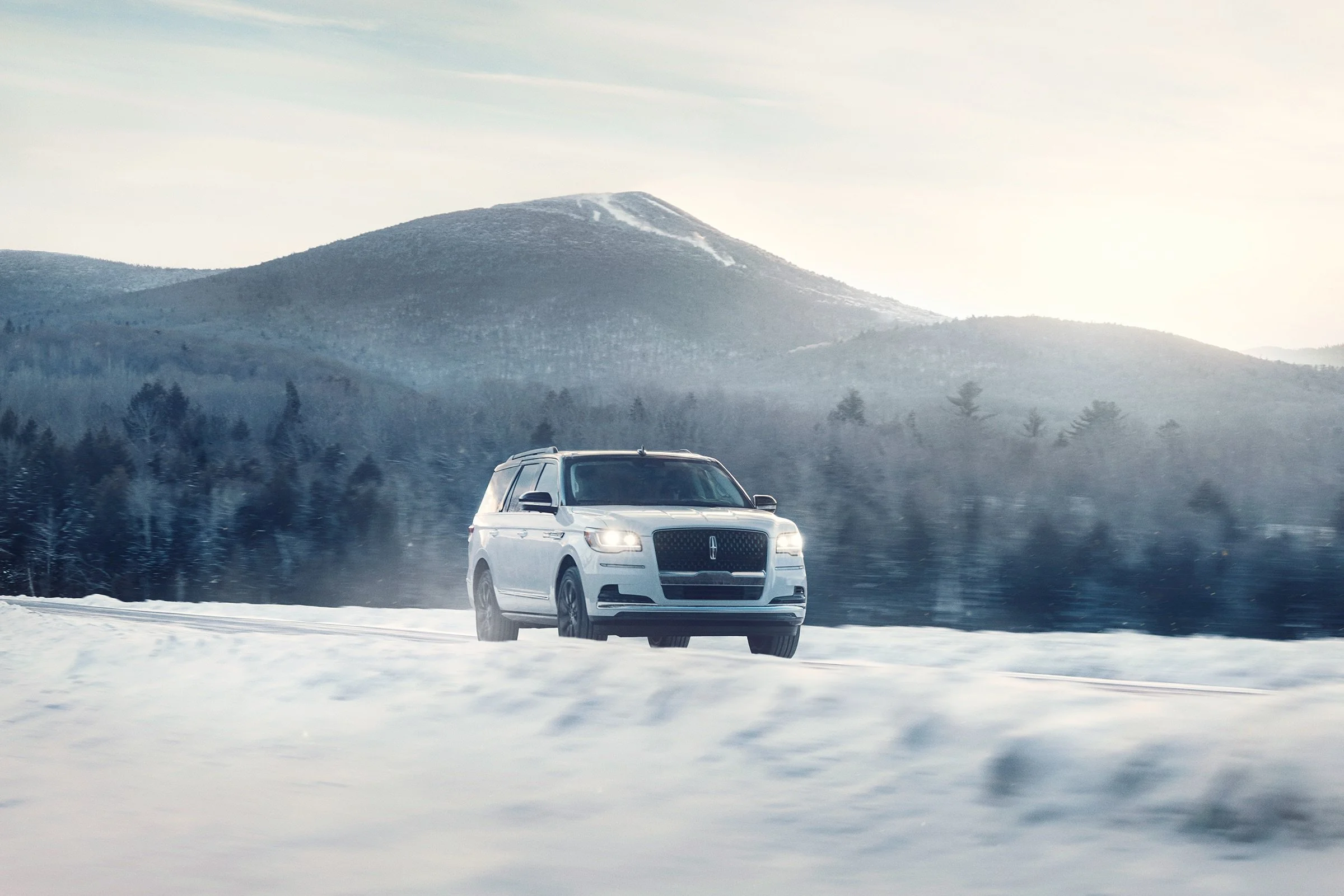 A white SUV driving through snow in a winter landscape with mountains and trees in the background.