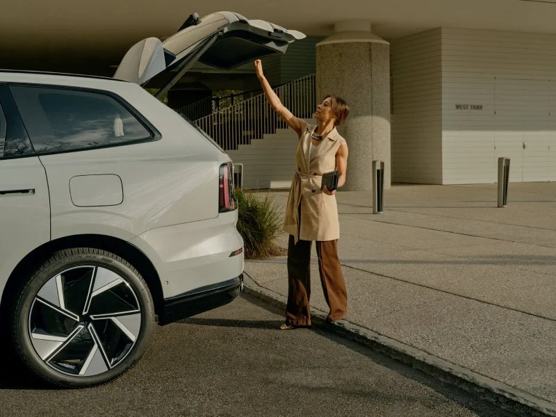 A woman is standing outside with her hand raised towards the open trunk of a white SUV.