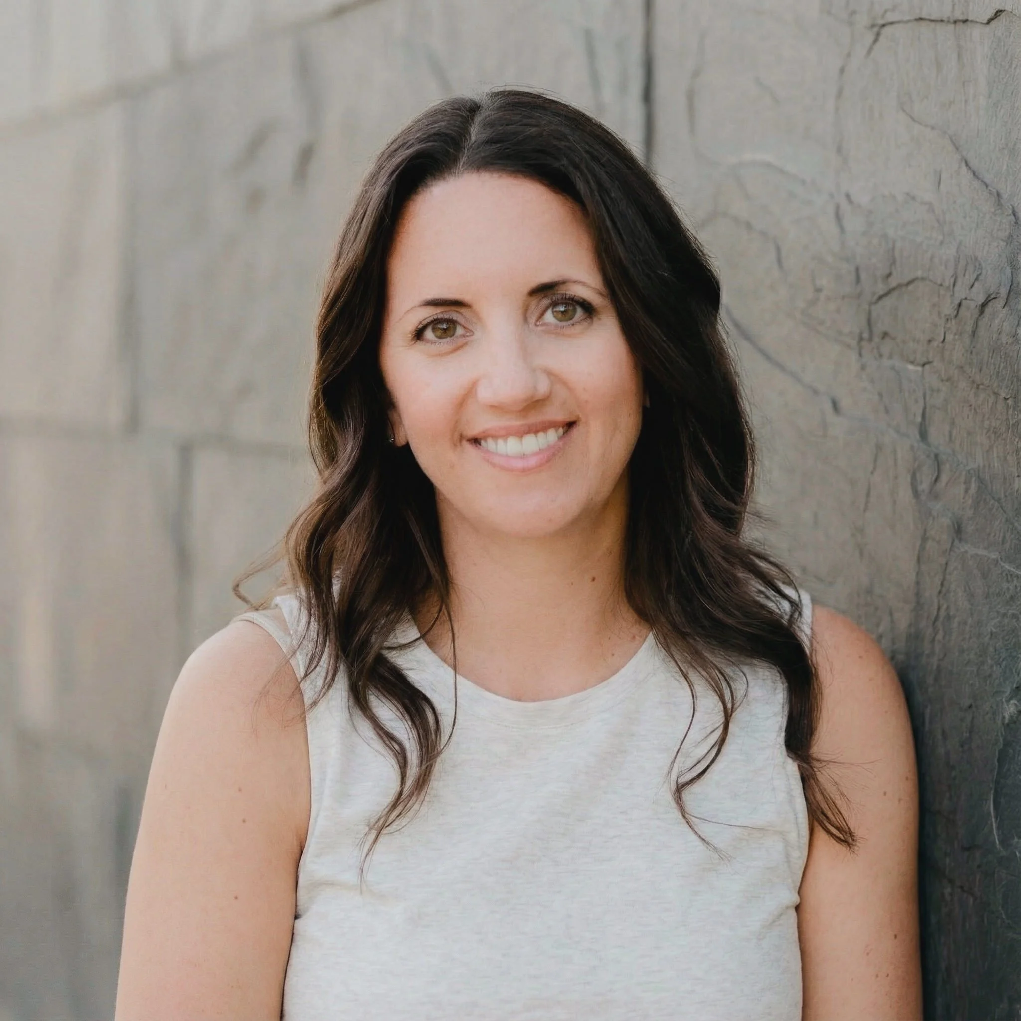 A woman with long dark hair and light skin smiling at the camera, standing against a stone wall.