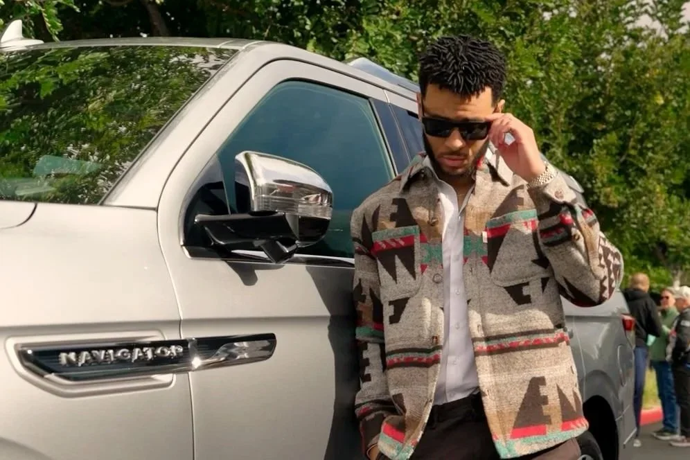 A man with dark, curly hair wearing sunglasses, a colorful patterned jacket, and a white shirt, standing next to a silver Lincoln Navigator SUV and adjusting his glasses outdoors.