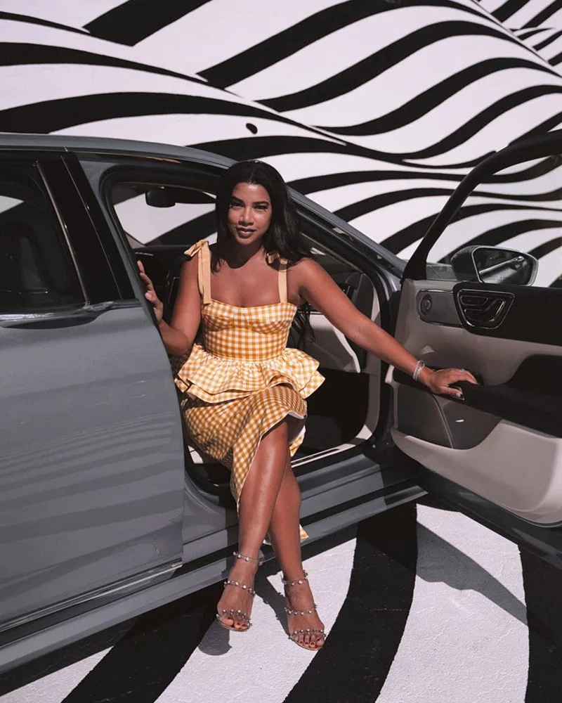 A woman in a yellow checkered dress sitting on the edge of a gray car with the driver's door open, in front of a black and white striped background.