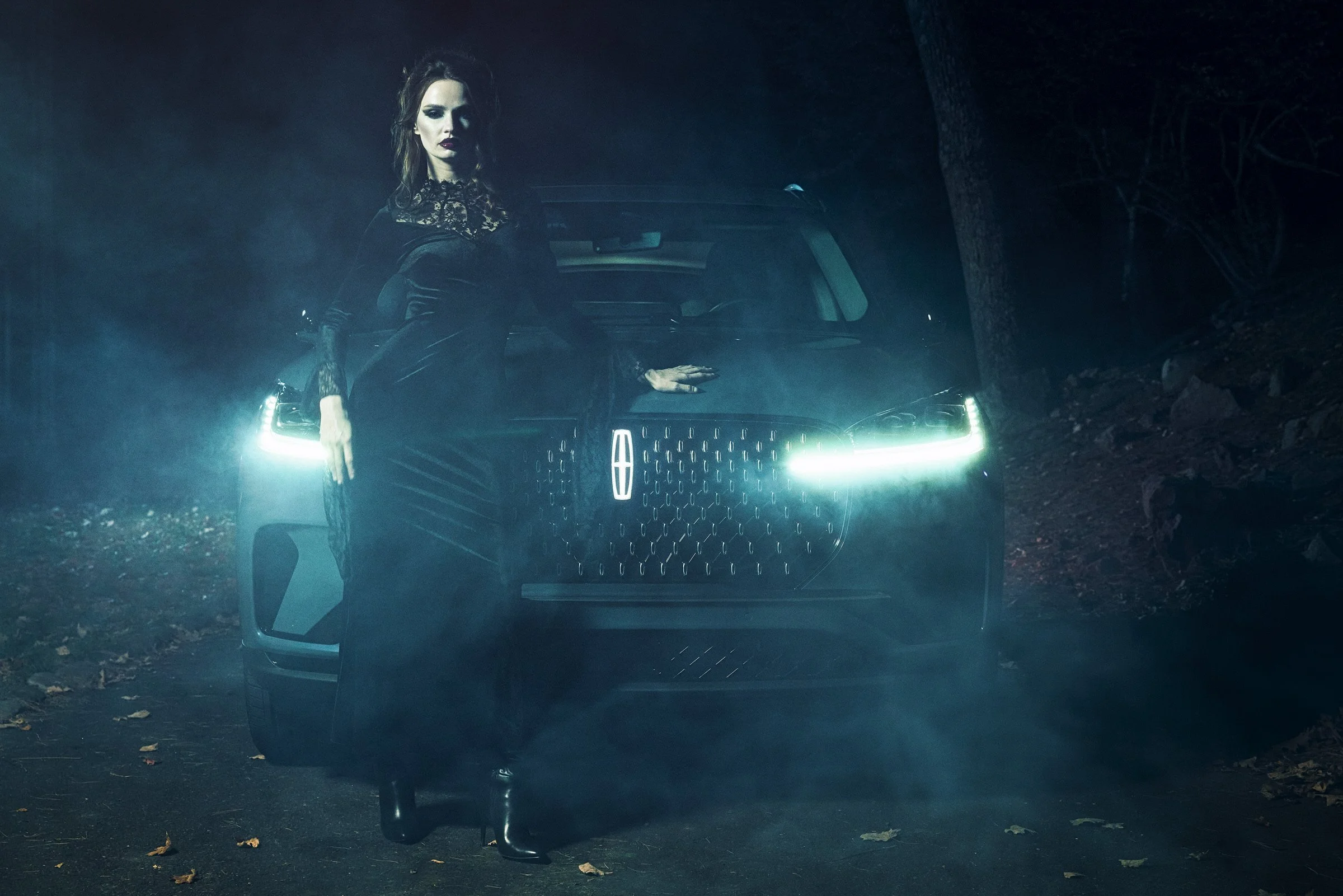 A woman dressed in black with dark makeup stands in front of a black car with illuminated headlights in a foggy, wooded area at night.