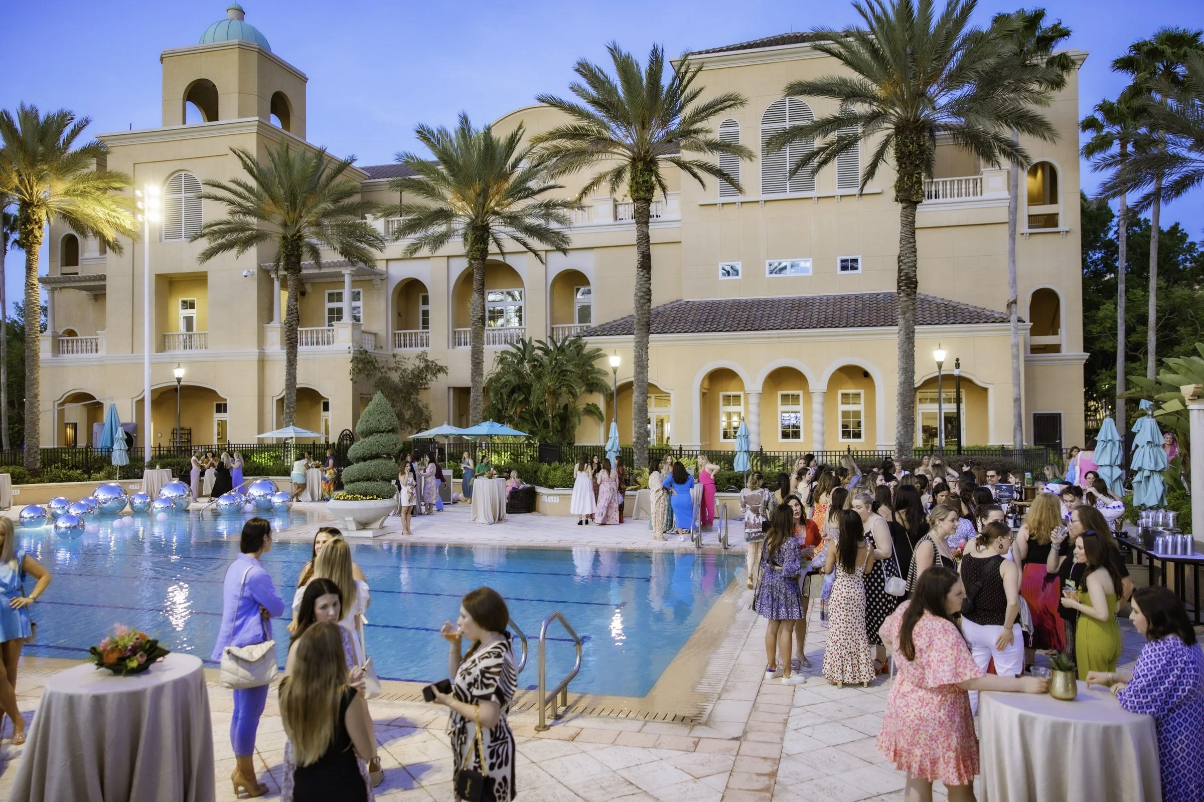 Poolside party at a luxurious hotel or resort with many guests mingling, palm trees, and a large beige building in the background during early evening.