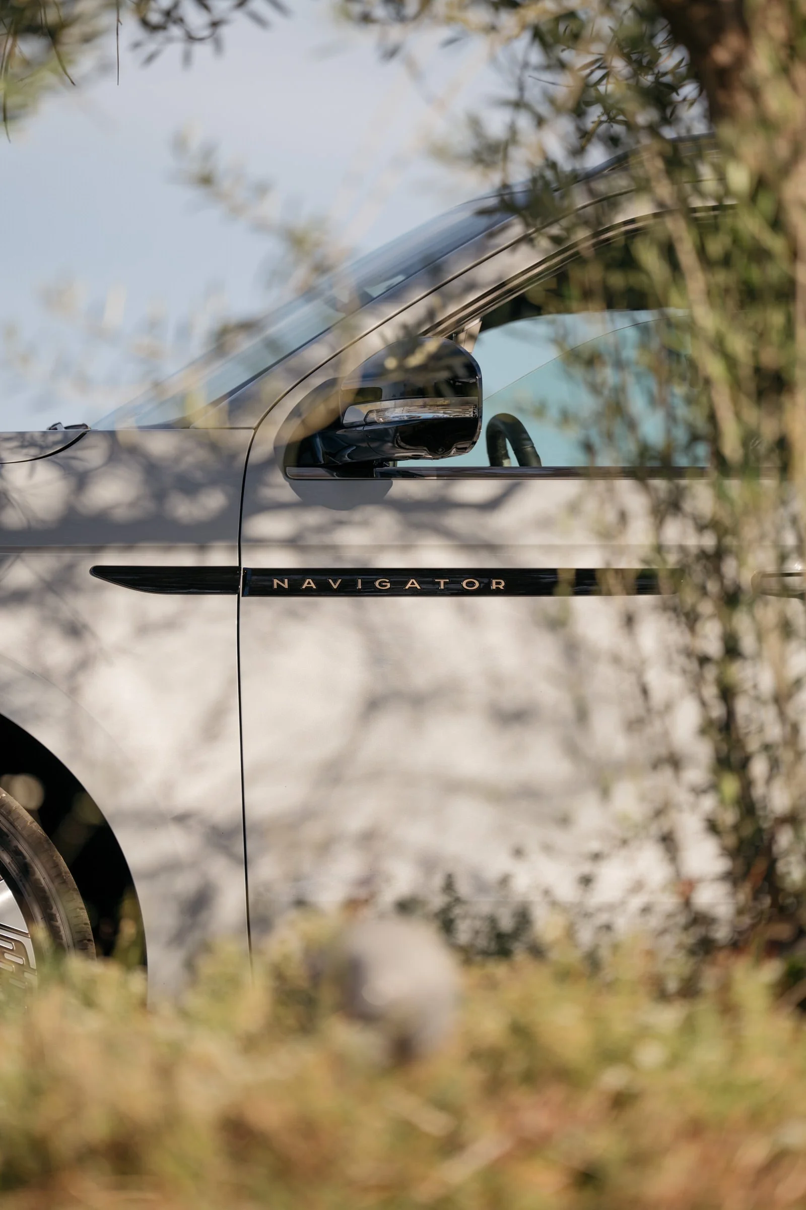 Part of a silver vehicle with the word 'NAVIGATOR' on the side, partially obscured by plants and trees.