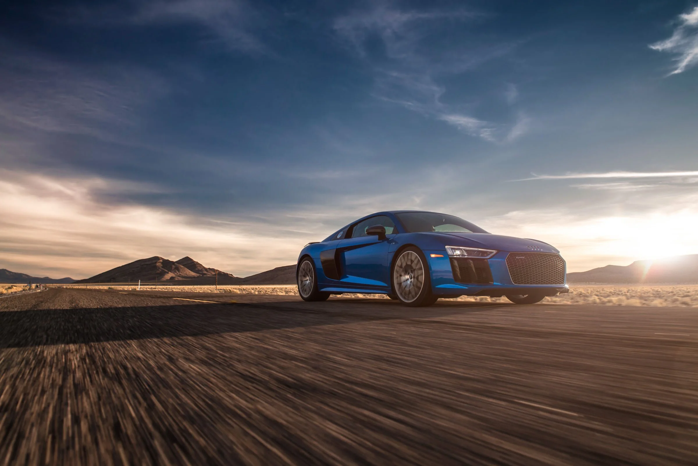 A blue sports car driving on an open road in a desert landscape during sunset with mountains in the background.