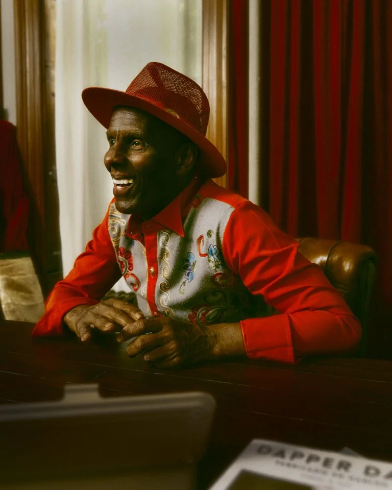A man smiling and wearing a red hat with a wide brim and a red jacket and vest with colorful embroidery, sitting at a table.
