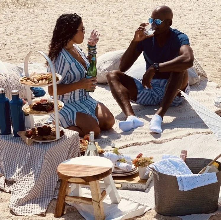 Man and woman having a picnic on the beach with food, drinks, and beach accessories.