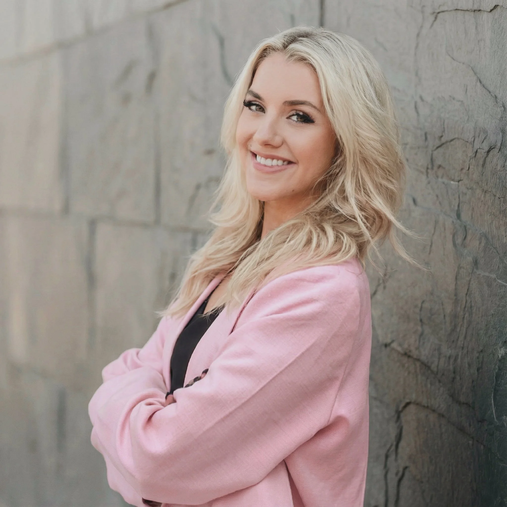 A young woman with blonde, wavy hair smiling and standing with arms crossed against a stone wall, wearing a pink blazer over a black top.