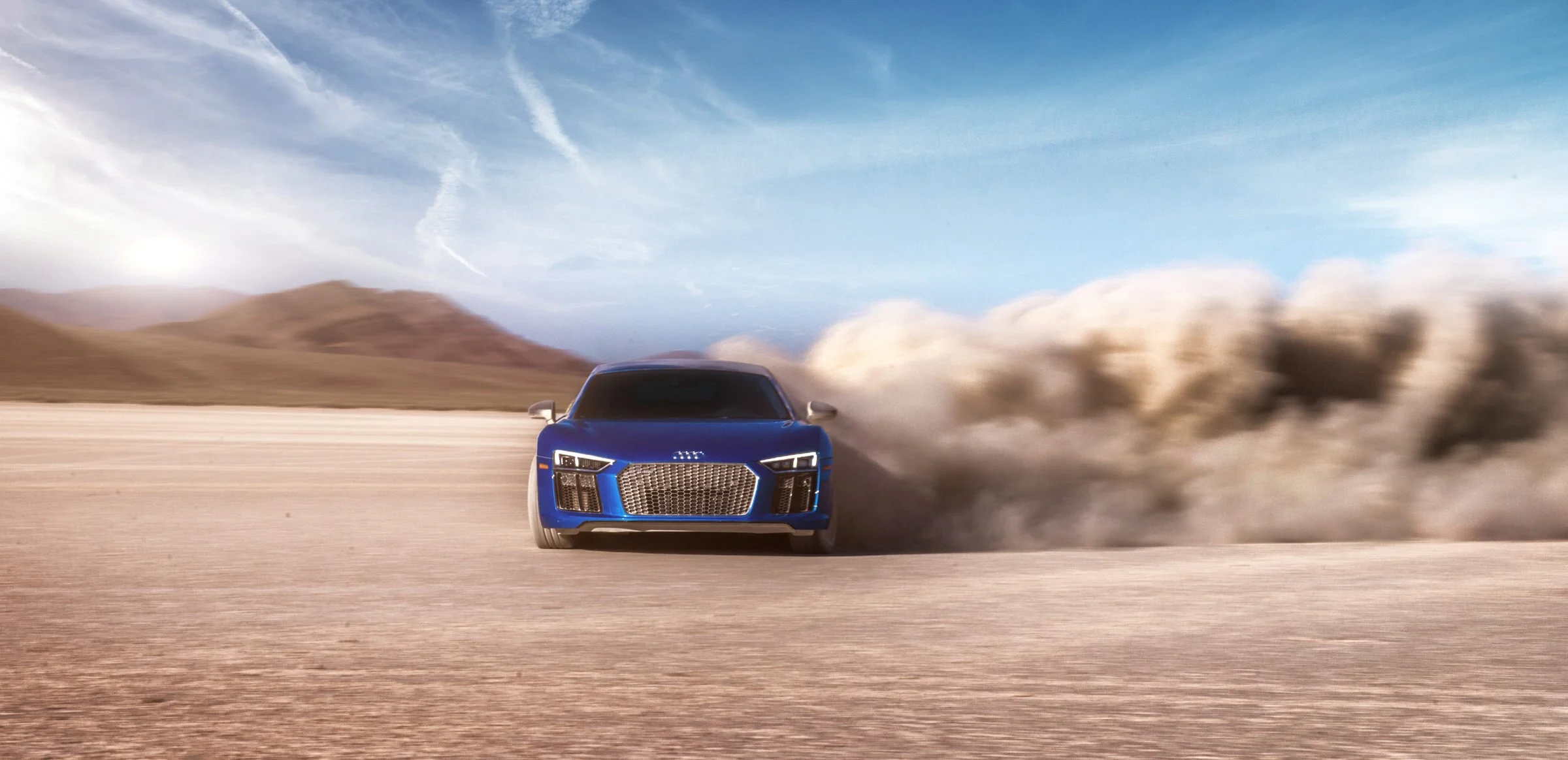 A blue sports car driving across a desert landscape, kicking up dust with mountains in the background and a partly cloudy sky overhead.