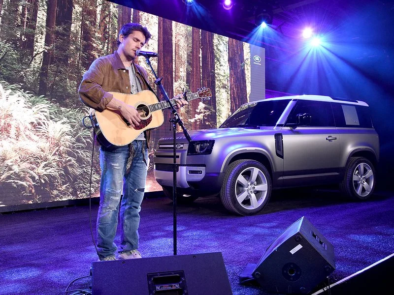 A man playing an acoustic guitar and singing into a microphone on stage, with an SUV vehicle and forest background projection behind him, and stage lights casting blue and purple hues.
