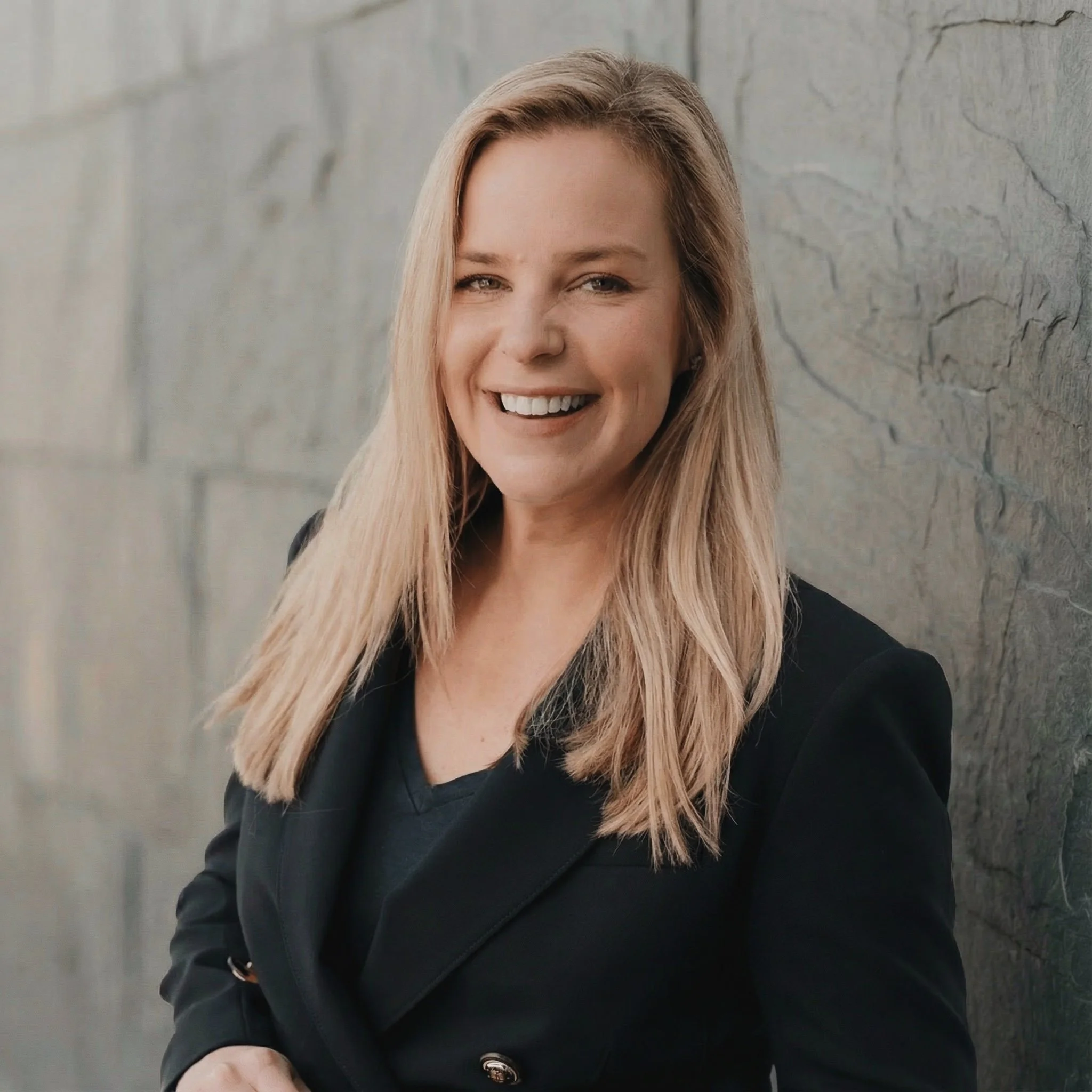 A woman with long blonde hair, smiling, wearing a black blazer, standing against a textured stone wall.