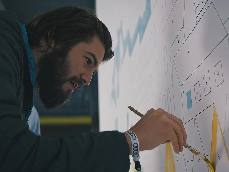 A man with long brown hair and a beard drawing or writing on a large whiteboard or wall with a marker, with charts and diagrams in the background.