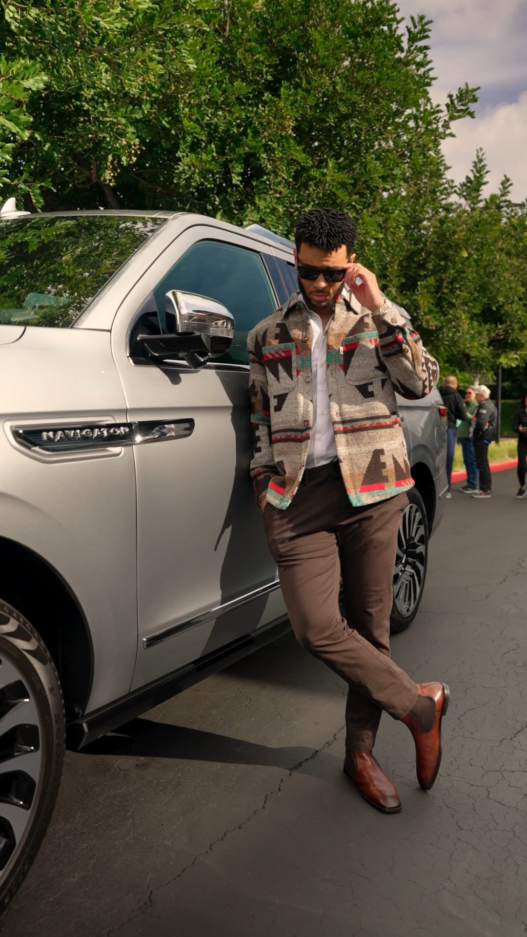 A man wearing sunglasses, a patterned jacket, white shirt, brown pants, and brown leather boots leans against a silver vehicle labeled 'Navigator' while talking on his phone. There are trees and a group of people in the background.