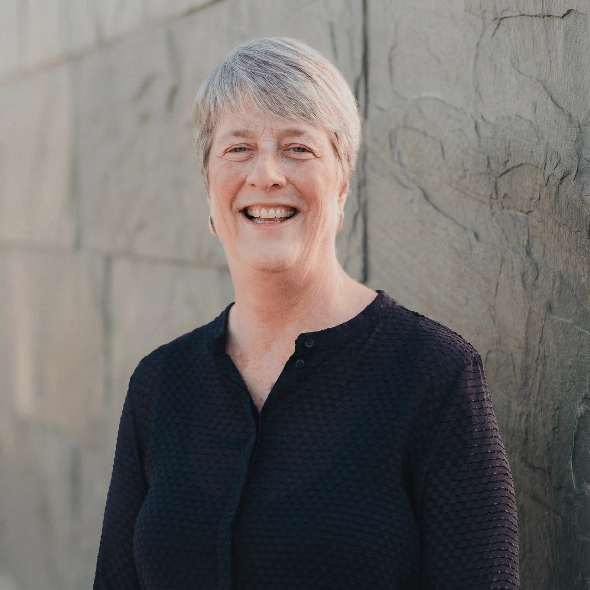 A smiling woman with short gray hair standing against a gray stone wall.