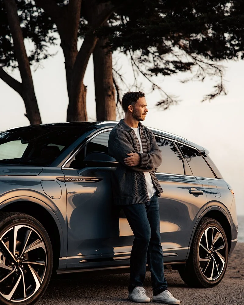 A man leaning against a blue luxury SUV parked outdoors near trees during sunset.