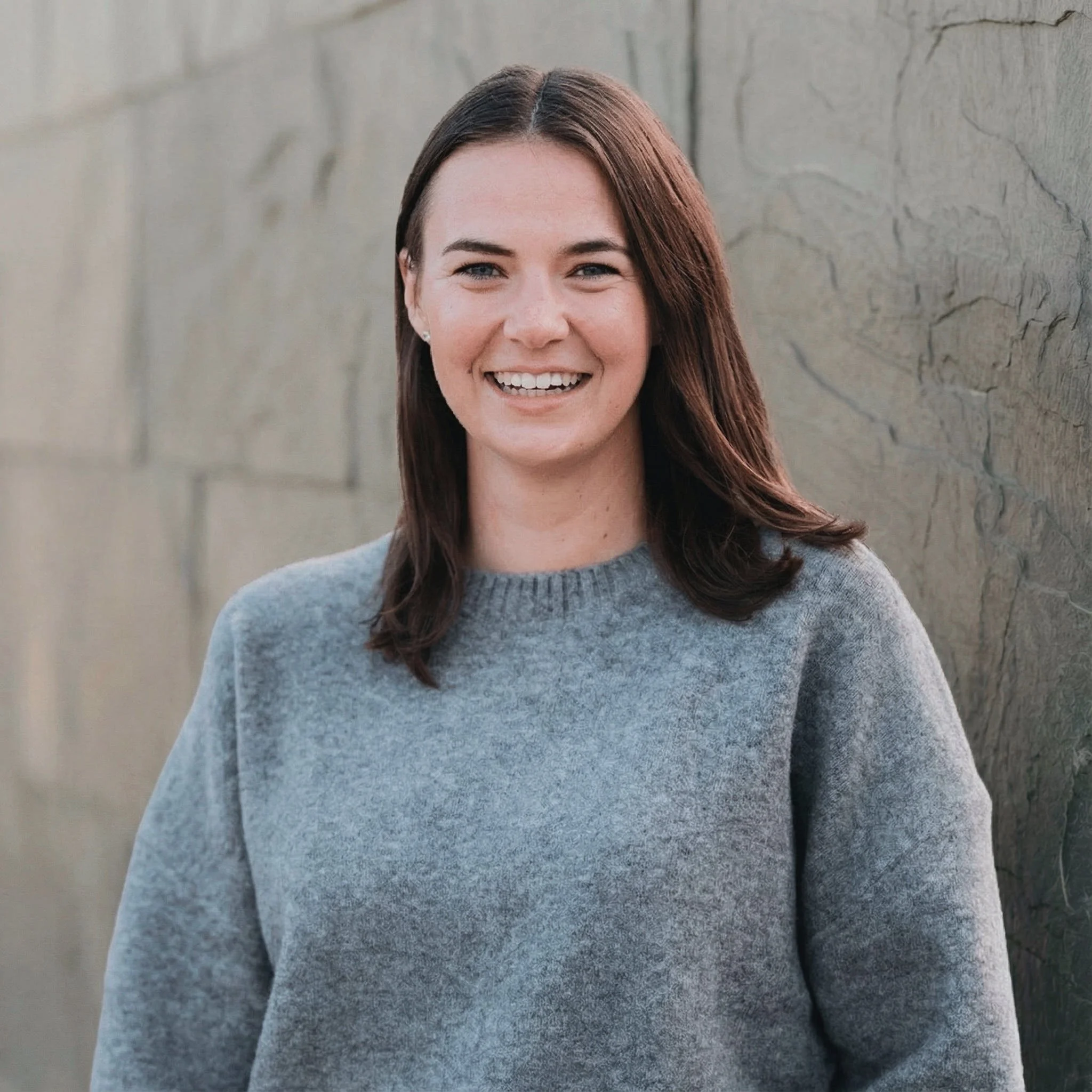 A woman with shoulder-length brown hair, smiling, wearing a gray sweater, standing against a textured gray wall.