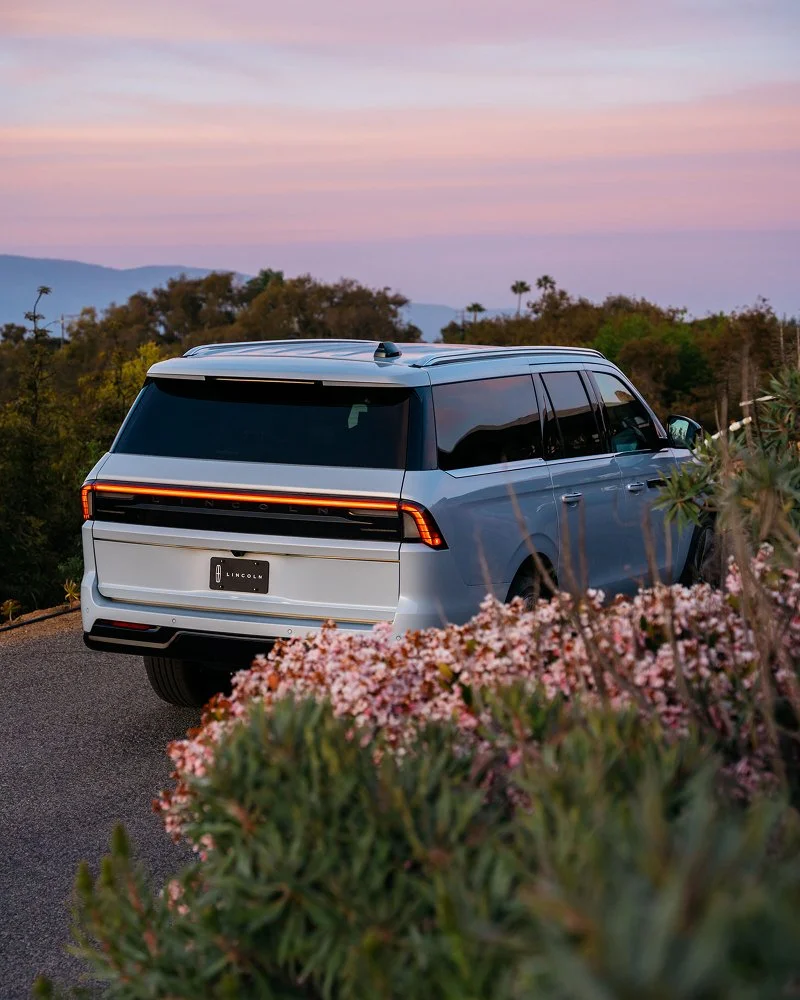 A white modern SUV parked on a scenic overlook during sunset with pink and purple sky, trees, and mountains in the background, and pink flowers in the foreground.