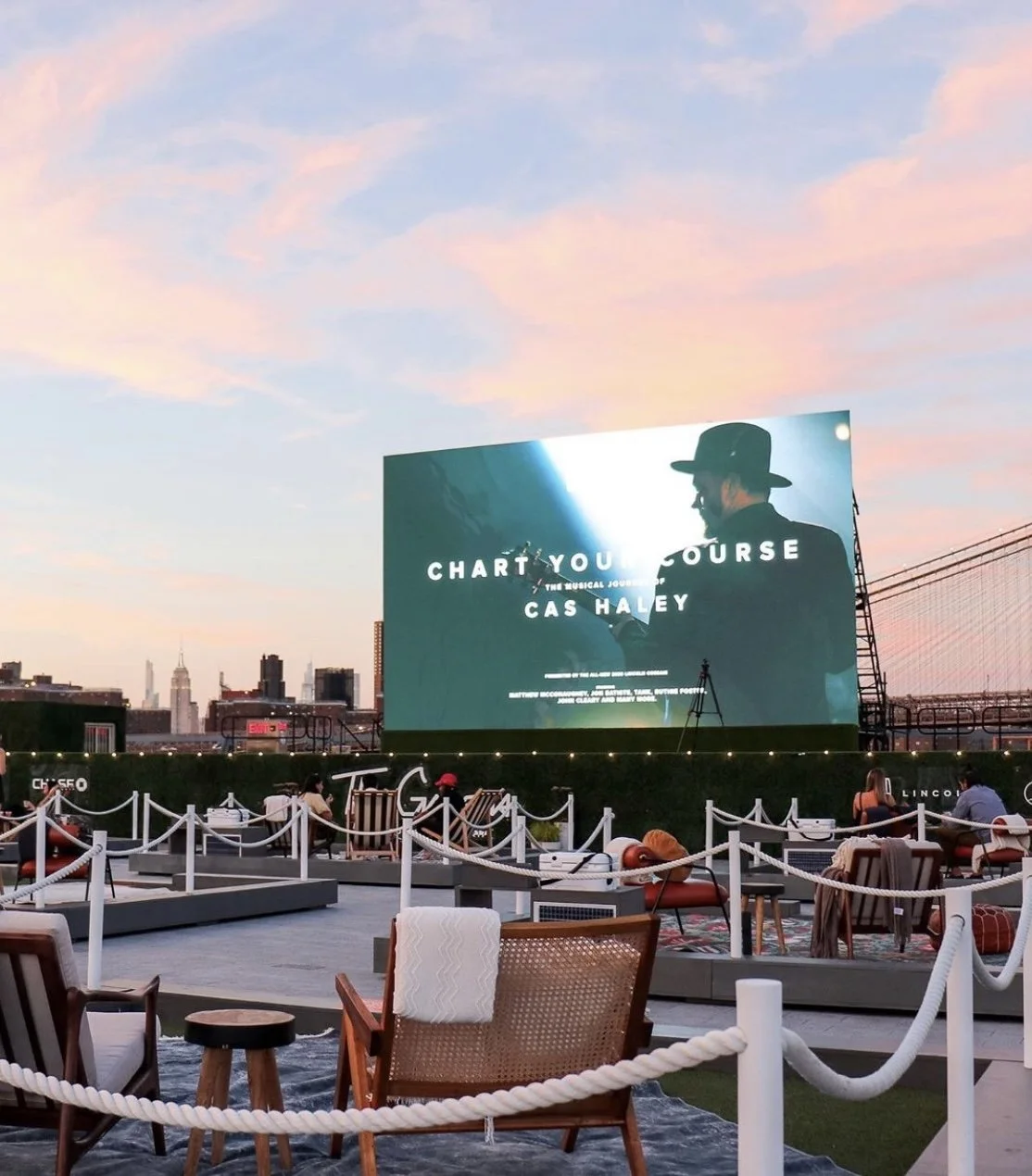 Outdoor rooftop cinema with a large screen displaying a promotional image for a musical called 'Charting Your Course' starring Cas Haley. There are chairs and small tables, some with people watching the screen at sunset. City skyline with tall buildings in the background.