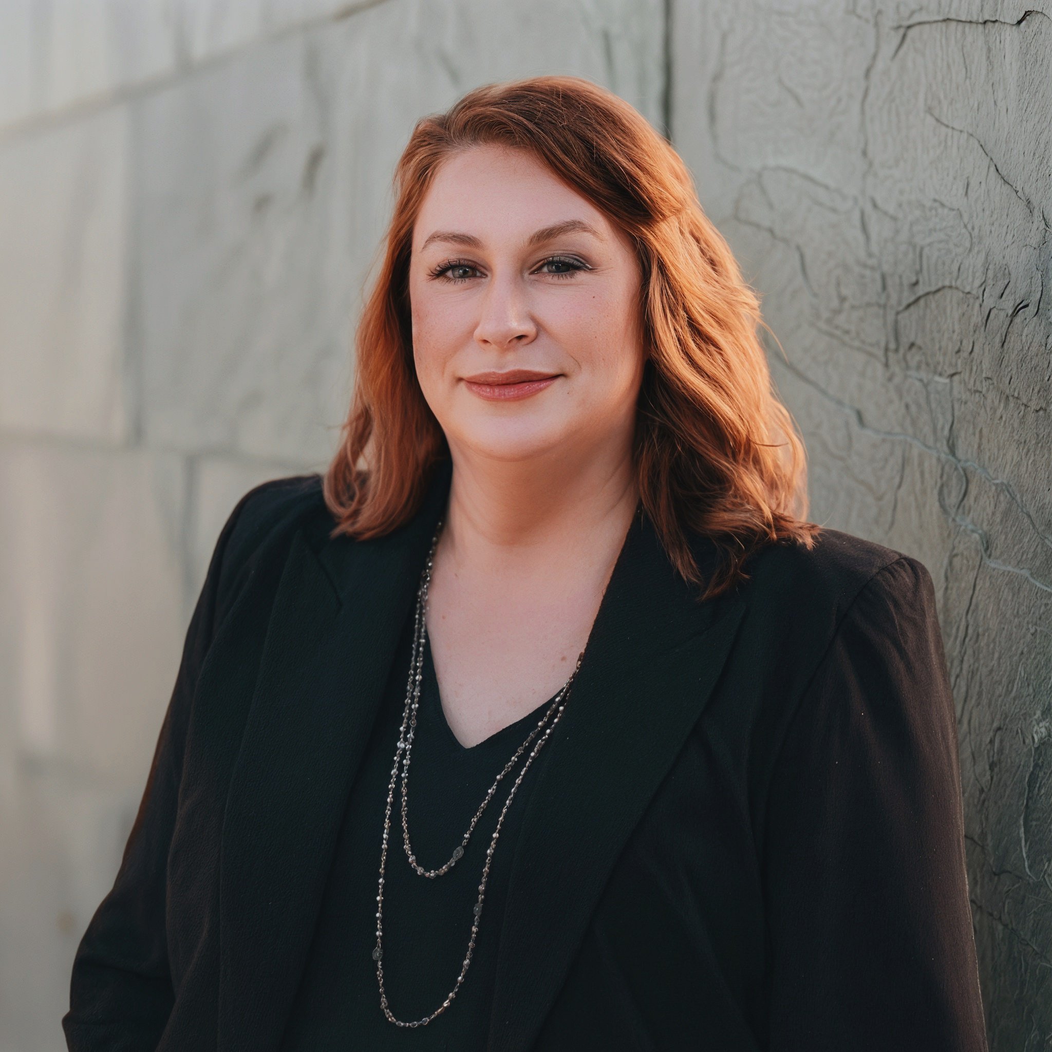 Portrait of a woman with shoulder-length red hair, wearing a black blazer and layered necklaces, standing against a textured wall.