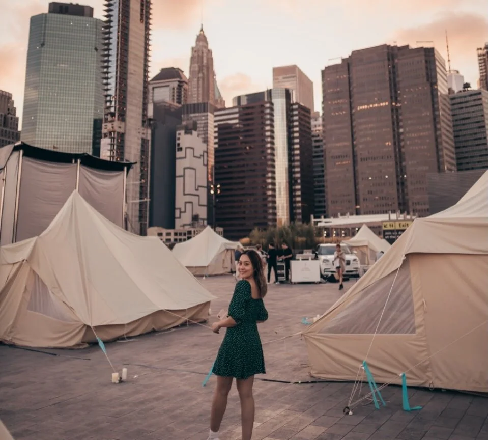 A woman smiling in a green dress with white polka dots standing among beige tents on a city parking lot at sunset, with skyscrapers in the background.