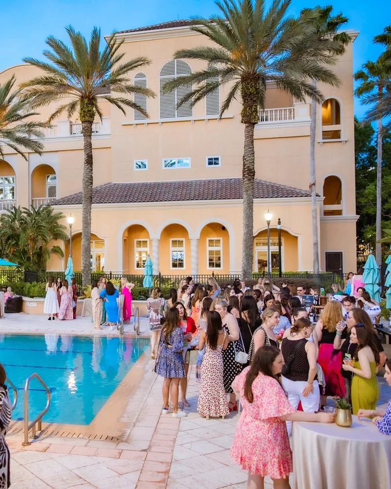 A poolside party at a luxury hotel with people socializing, palm trees, and a large beige building in the background during early evening.