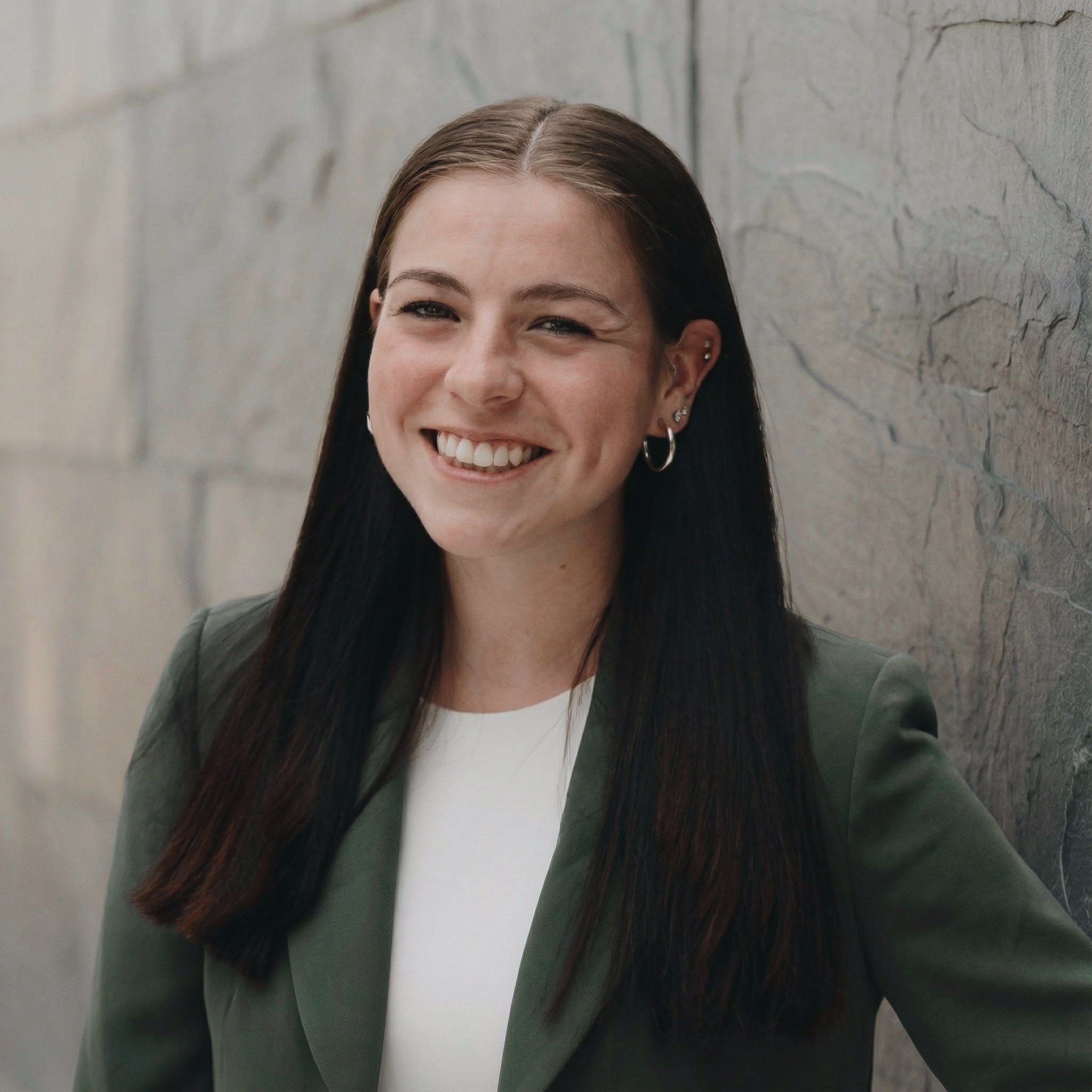 A woman with long dark hair, wearing a white top and green blazer, smiling and standing against a stone wall.