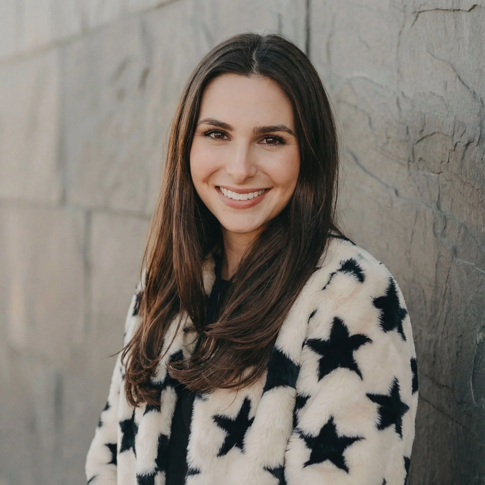 A young woman with long brown hair smiling, wearing a white faux fur coat with black stars, standing against a gray stone wall.
