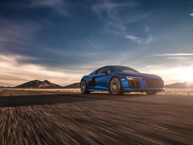 A blue sports car driving on an open desert road during sunset with mountains in the background.
