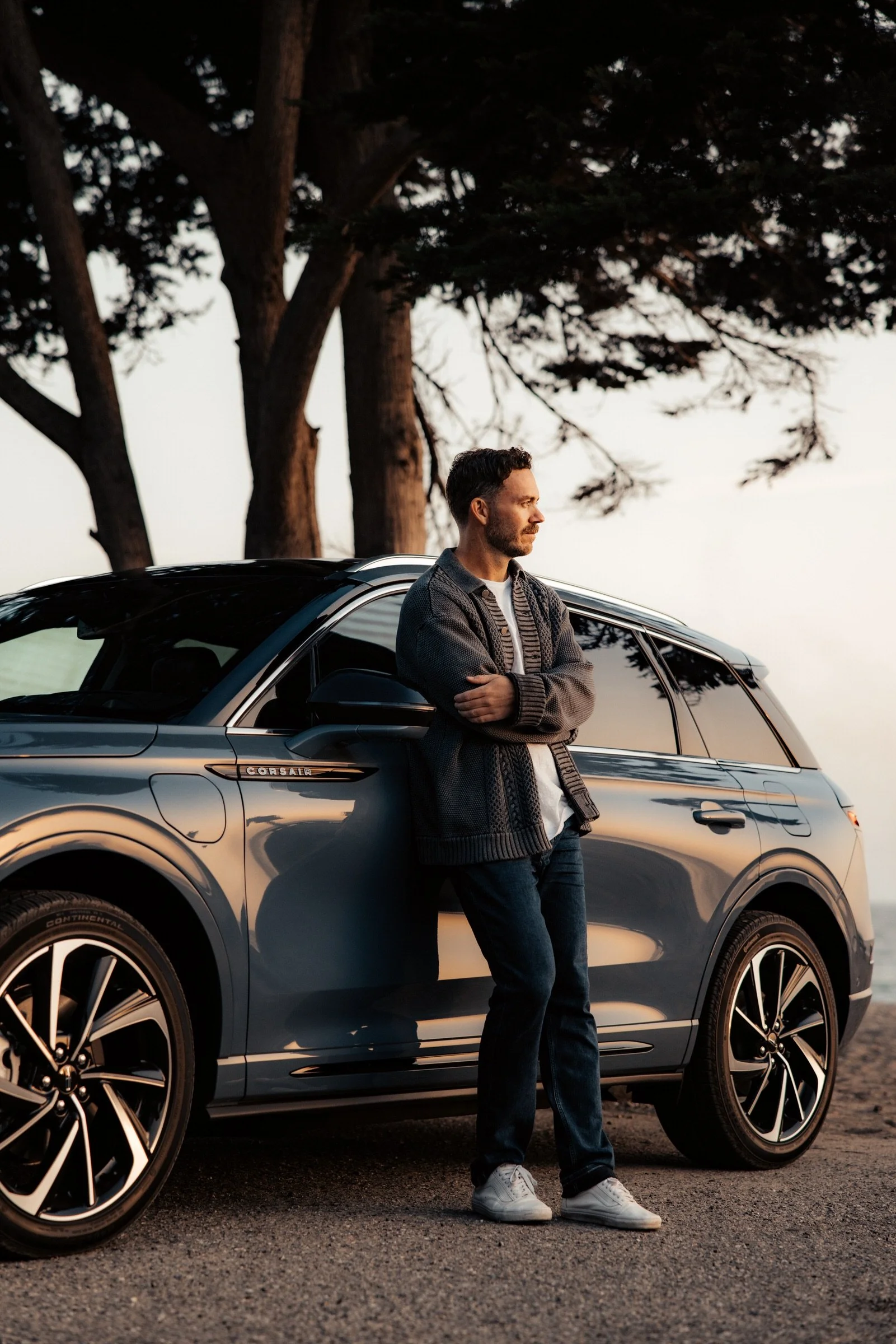 A man is standing next to a grey SUV with a 'Corsair' badge, leaning against the vehicle with arms crossed, looking into the distance with trees and a sunset in the background.