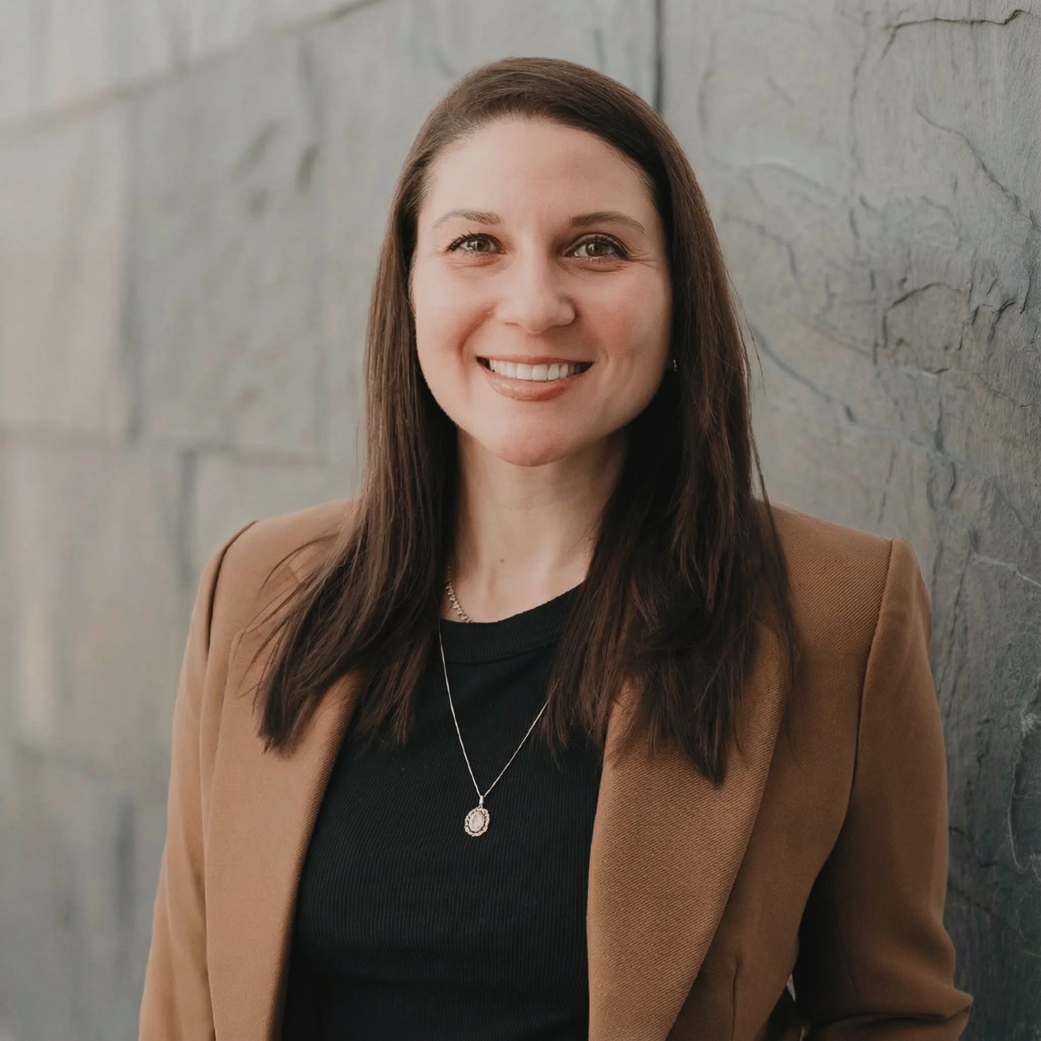 A smiling woman with shoulder-length brown hair, wearing a brown blazer over a black top, standing against a gray stone wall.