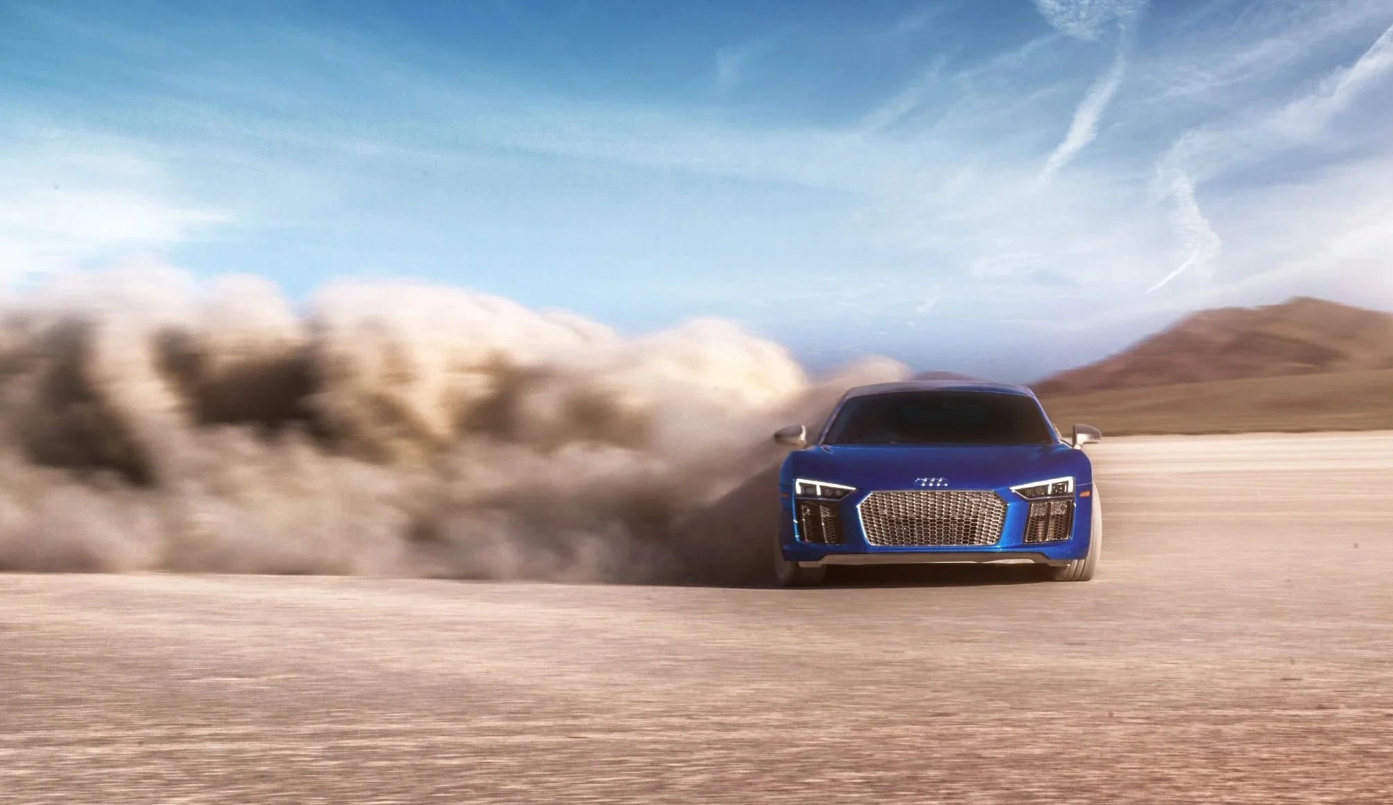 A blue car speeding on a desert road, kicking up dust with mountains in the background and a partly cloudy sky.