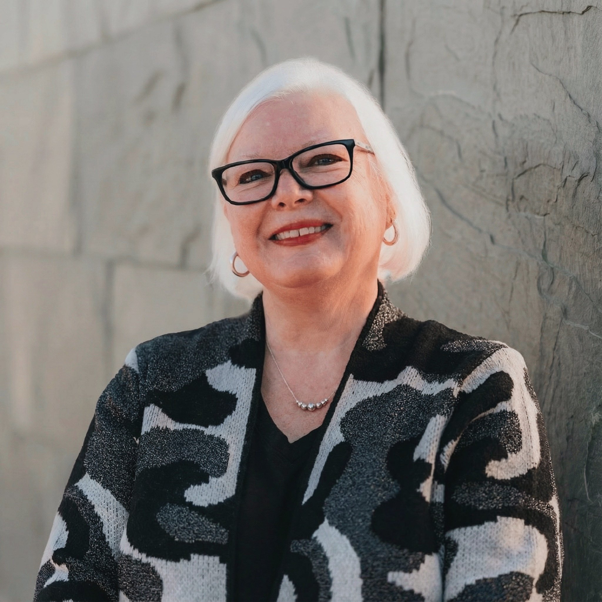 Senior woman with white hair, glasses, and jewelry, smiling outdoors near a stone wall.