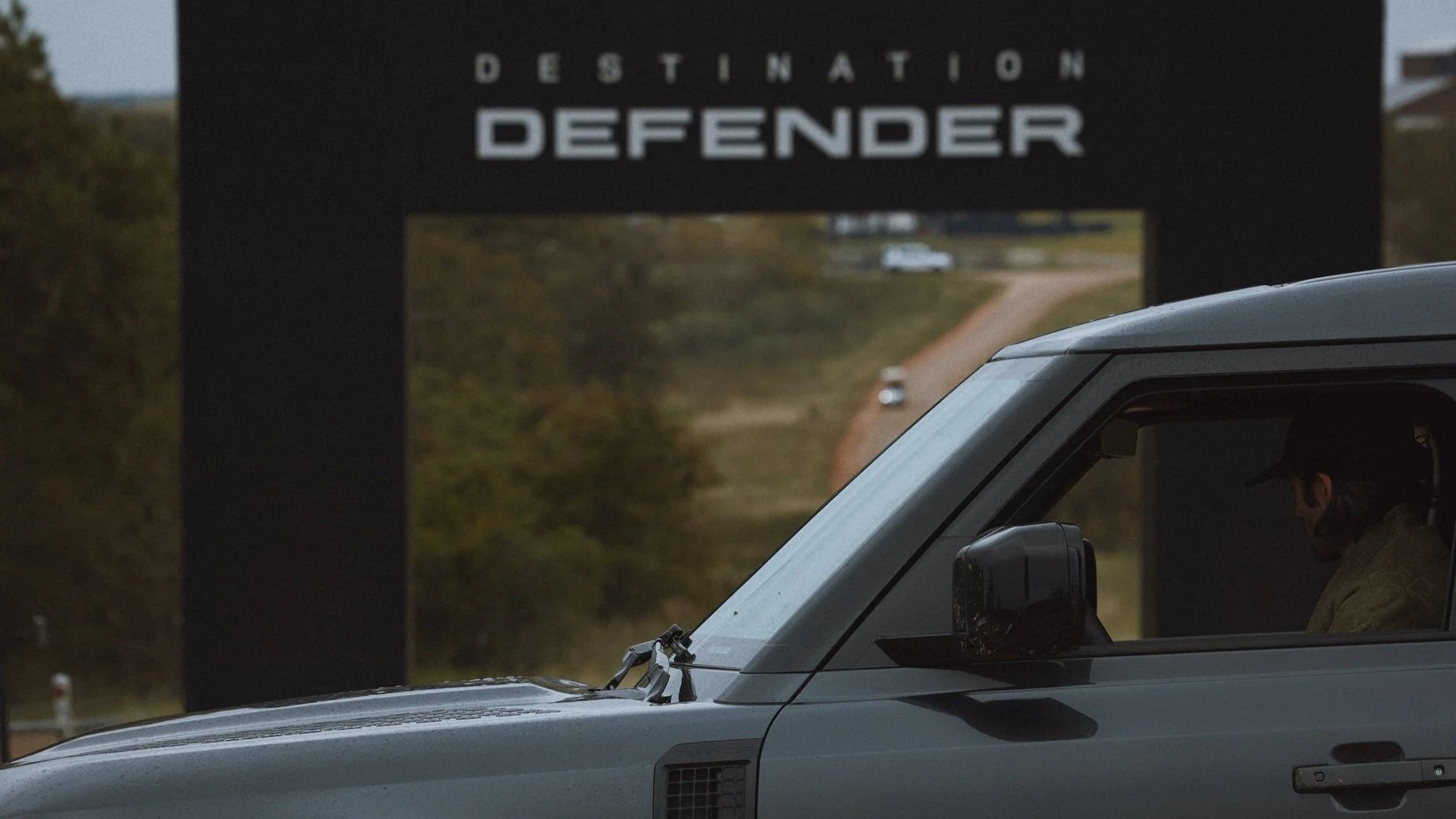 A gray vehicle with a driver wearing a face mask parked on the side of a dirt road leading towards a large sign that reads 'Destination Defender'.