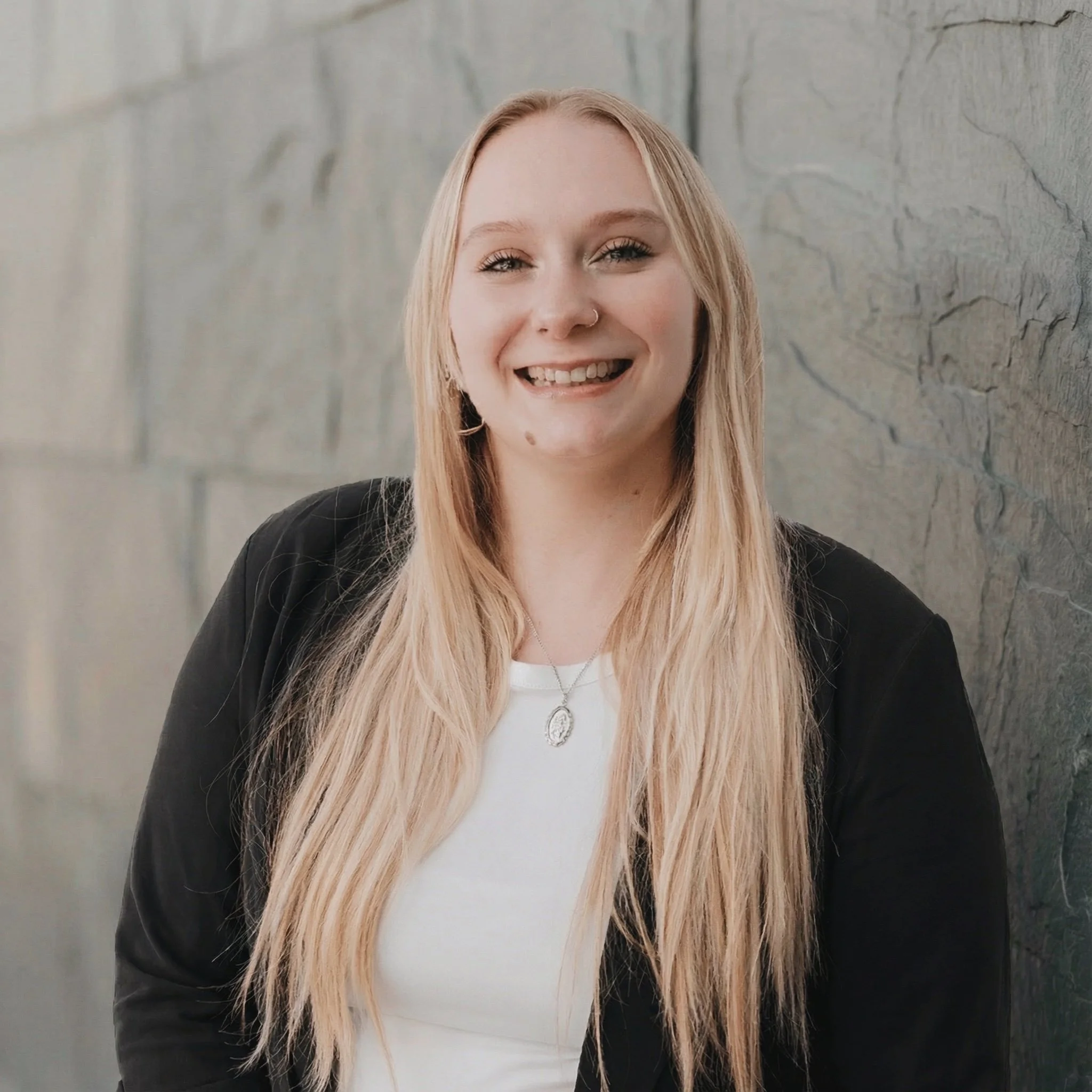A young woman with long blonde hair, smiling, wearing a black jacket over a white shirt, standing against a stone wall.
