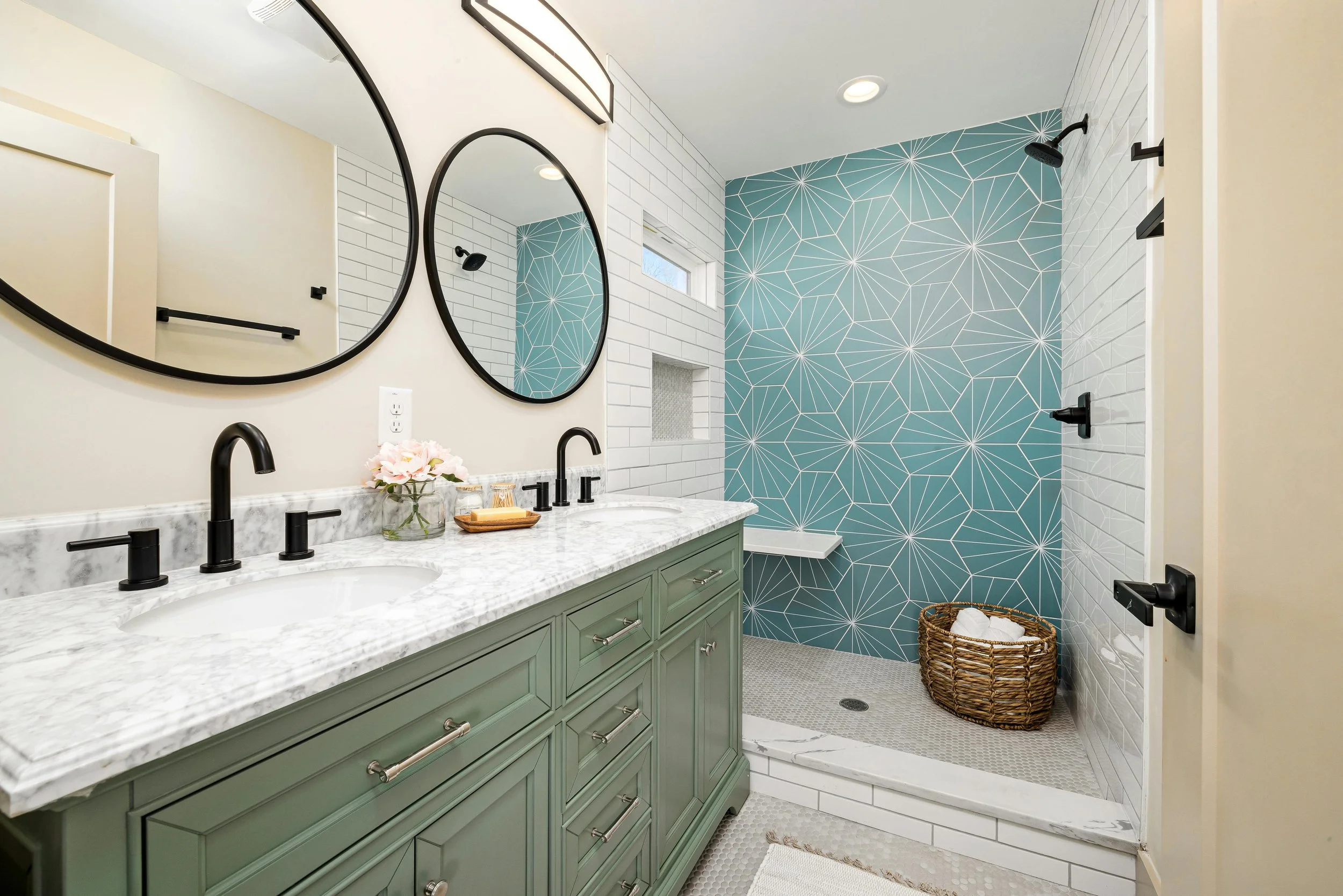 Modern bathroom with a white marble double vanity, black fixtures, and two round mirrors. A walk-in shower has geometric blue tile on one wall and white subway tile on the others, with black fixtures and a woven basket of towels.