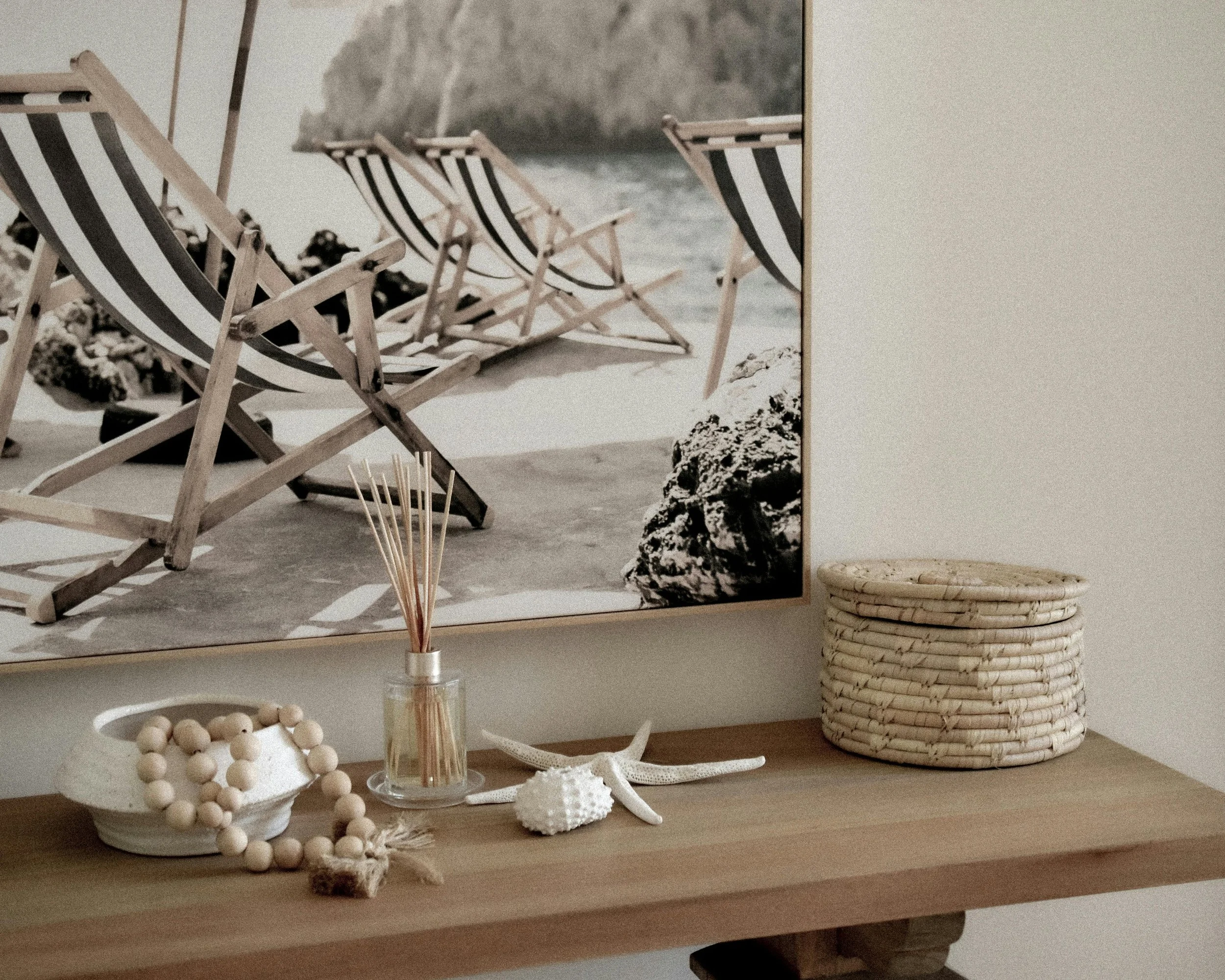 A wooden console table with decorative items, including a white shell-shaped dish with a beaded bracelet, a glass diffuser with reed sticks, a starfish, a seashell, and a woven basket. A framed photograph of striped beach chairs on sand with rocks and water is hung on the wall above the table.