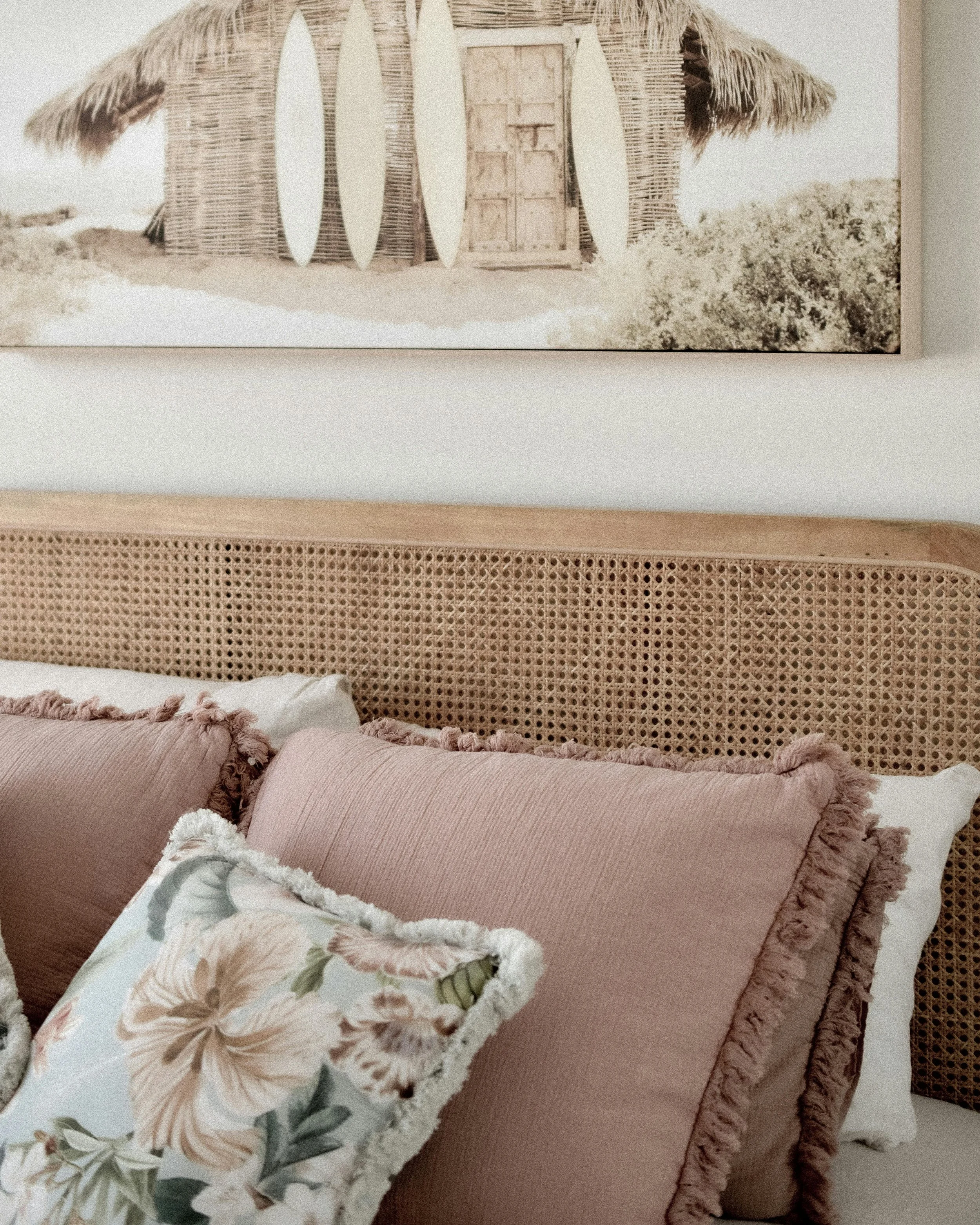 A bedroom with a rattan headboard, decorated with pink and floral pillows, and a painting of a beach hut with surfboards above it hanging on the wall.