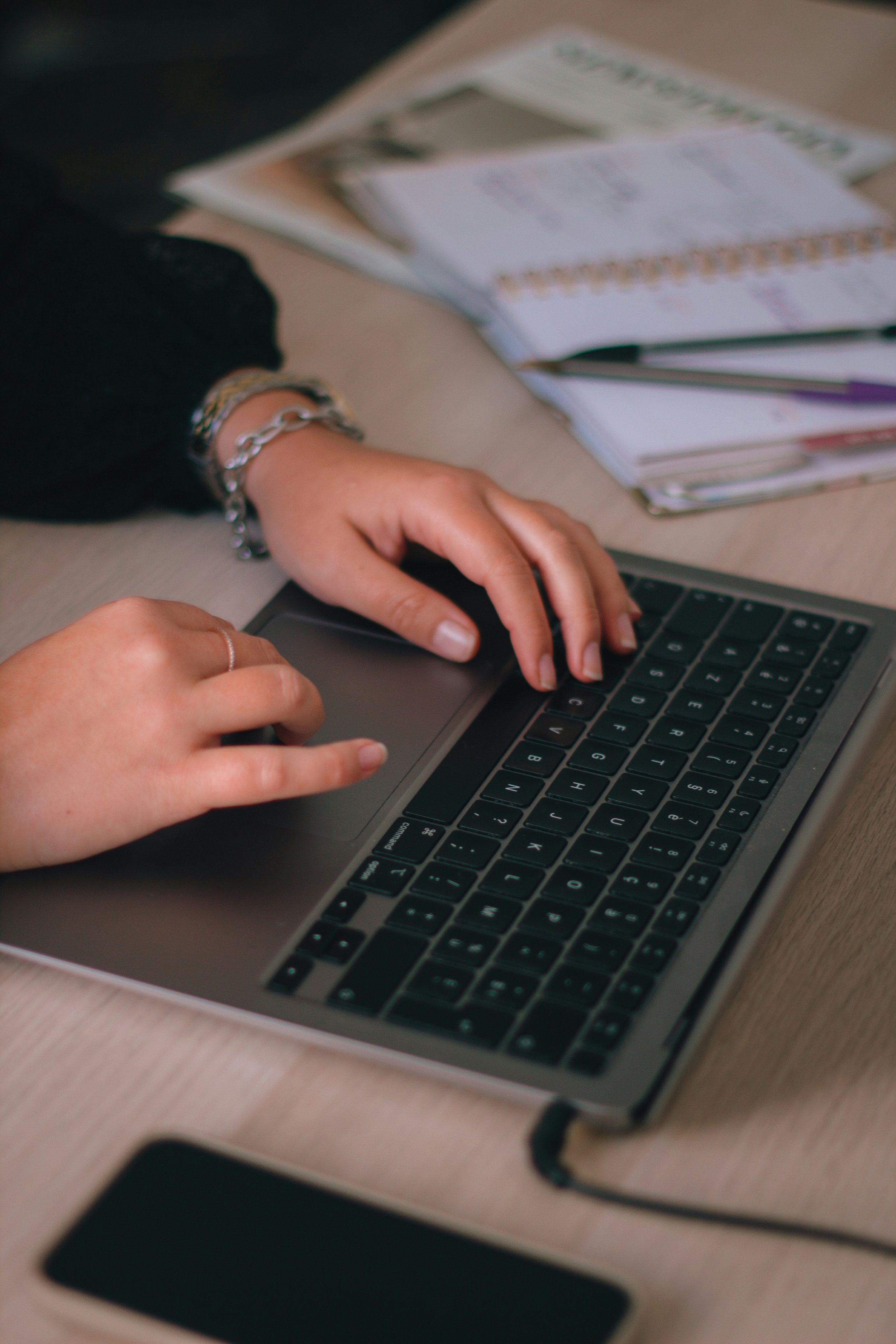A person type on a laptop keyboard, with documents and magazines on a desk.