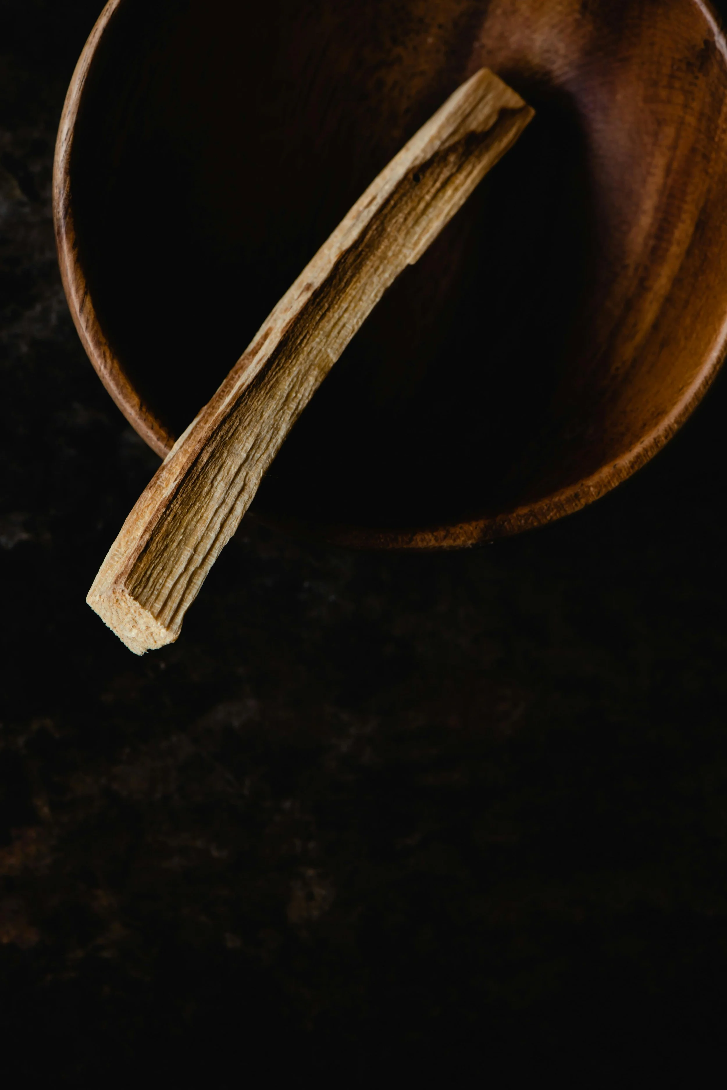 A wooden stick resting inside a dark wooden bowl on a black surface.