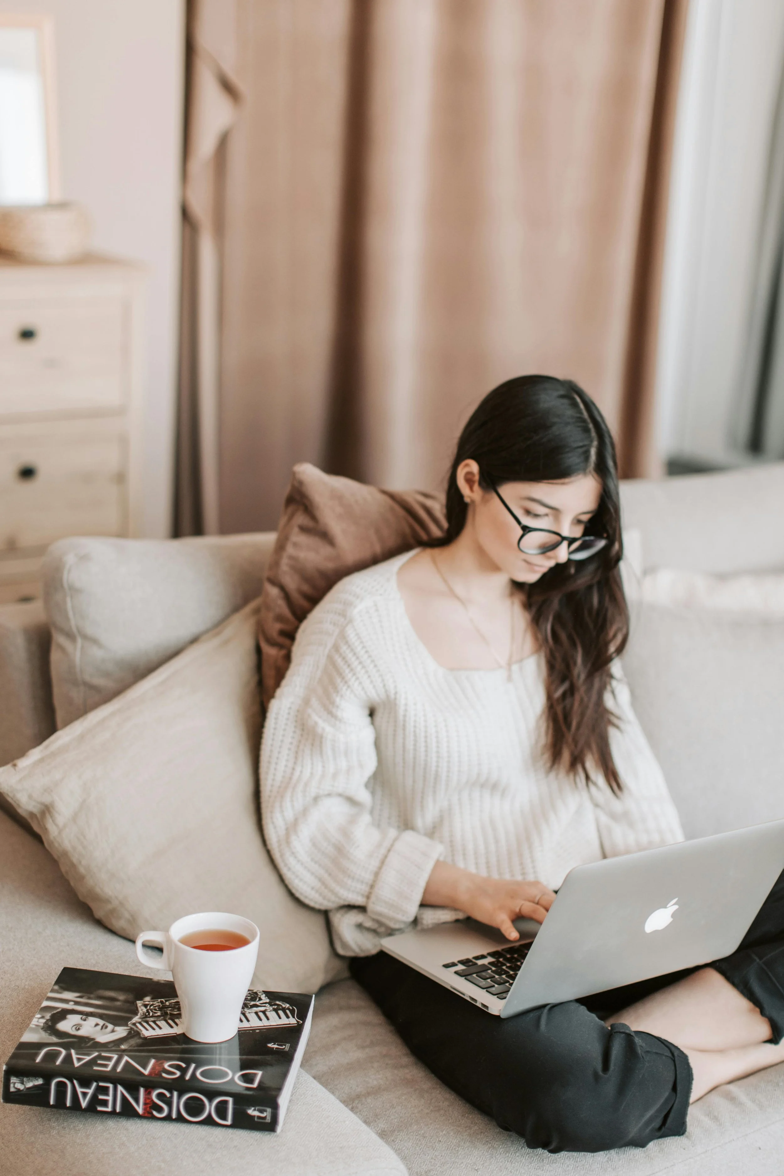 A woman with long dark hair and glasses working on a MacBook laptop on a beige couch, with a cup of tea and a fashion magazine nearby.