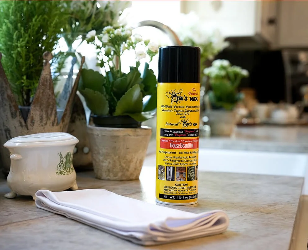 A can of bee's wax furniture polish on a kitchen countertop, with a white cloth and potted plants in the background.