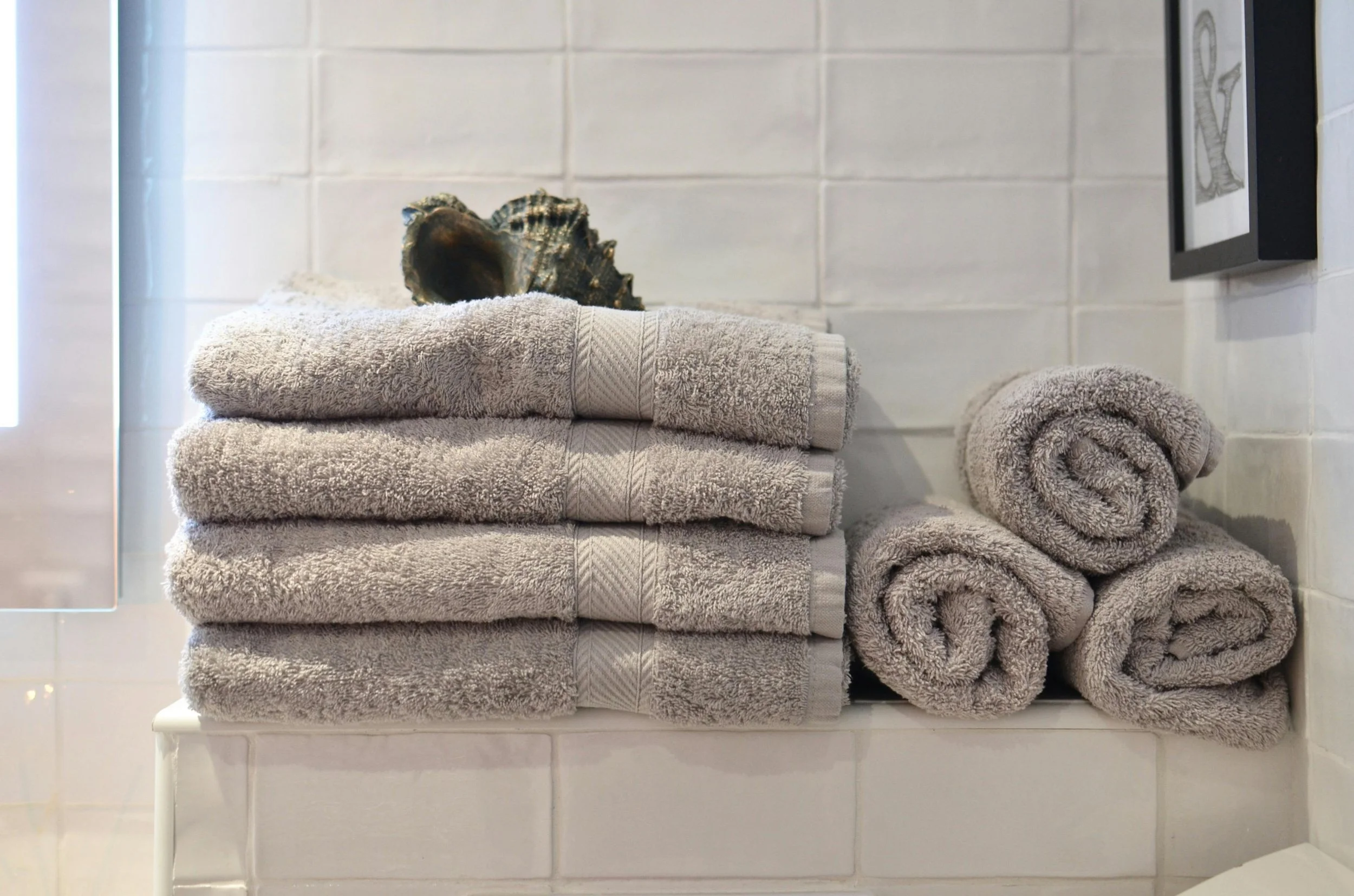 Stacked beige towels and a shell on a bathroom shelf against a white tiled wall.