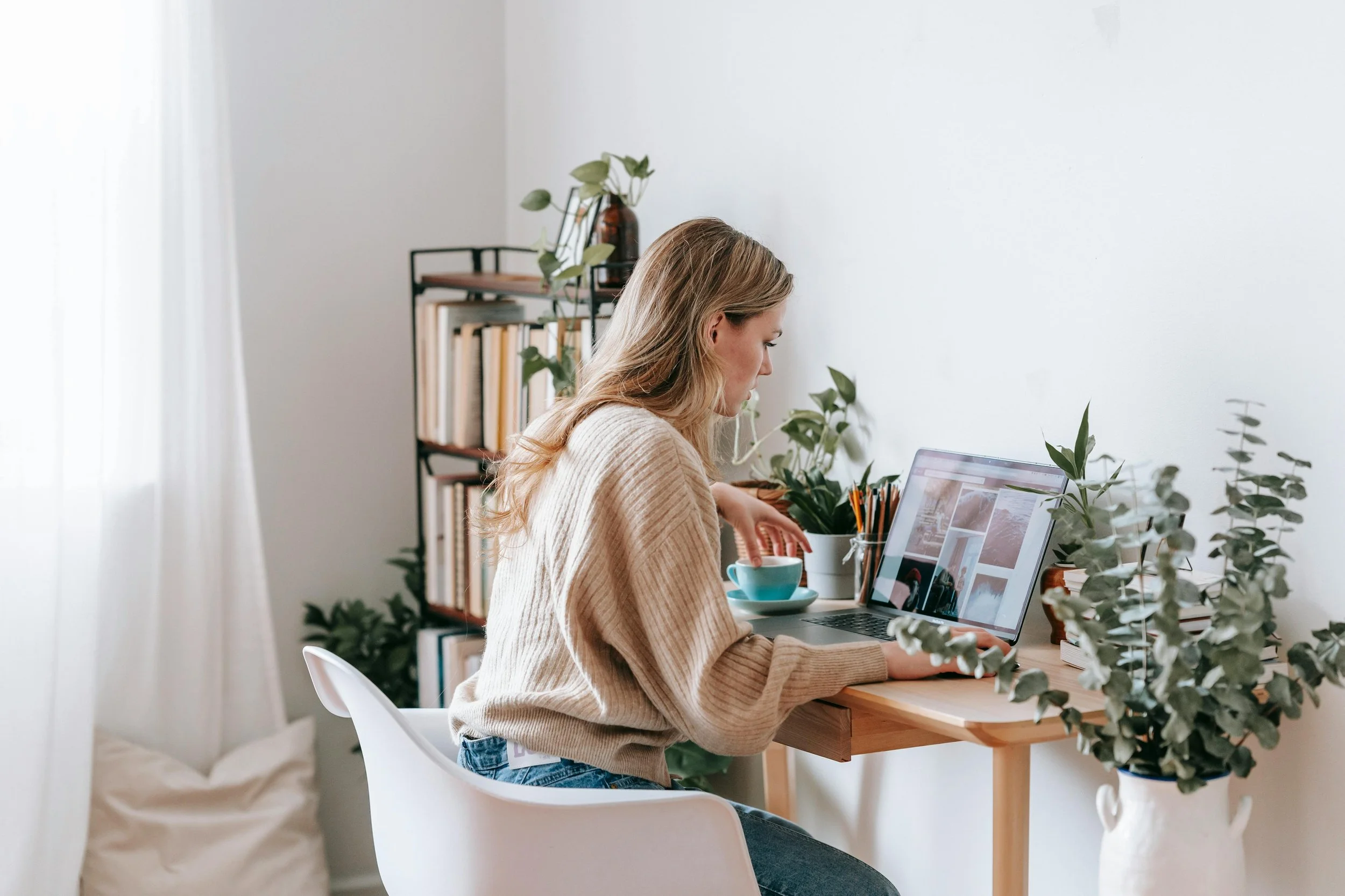 A woman sitting at a wooden desk working on a laptop, surrounded by green plants and a bookshelf.