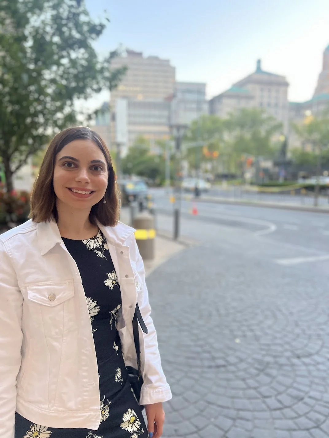 A woman with shoulder-length brown hair smiling outdoors on a city street, wearing a white jacket and a black dress with white floral patterns, with a cityscape of tall buildings and trees in the background.