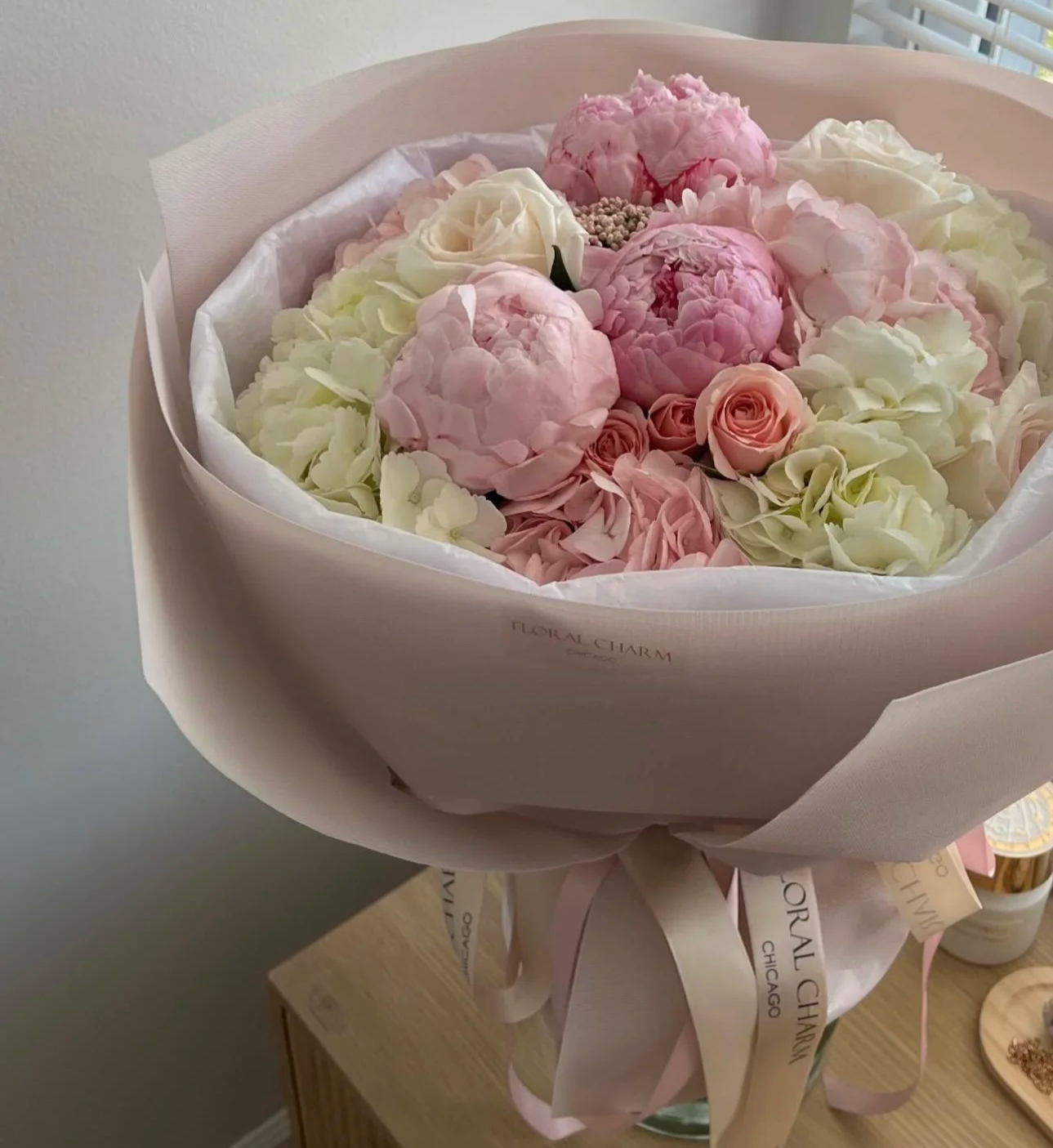 A large bouquet of pink and white flowers, including peonies and roses, wrapped in white and pink paper, with a ribbon that says 'Floral Charm Chicago'.