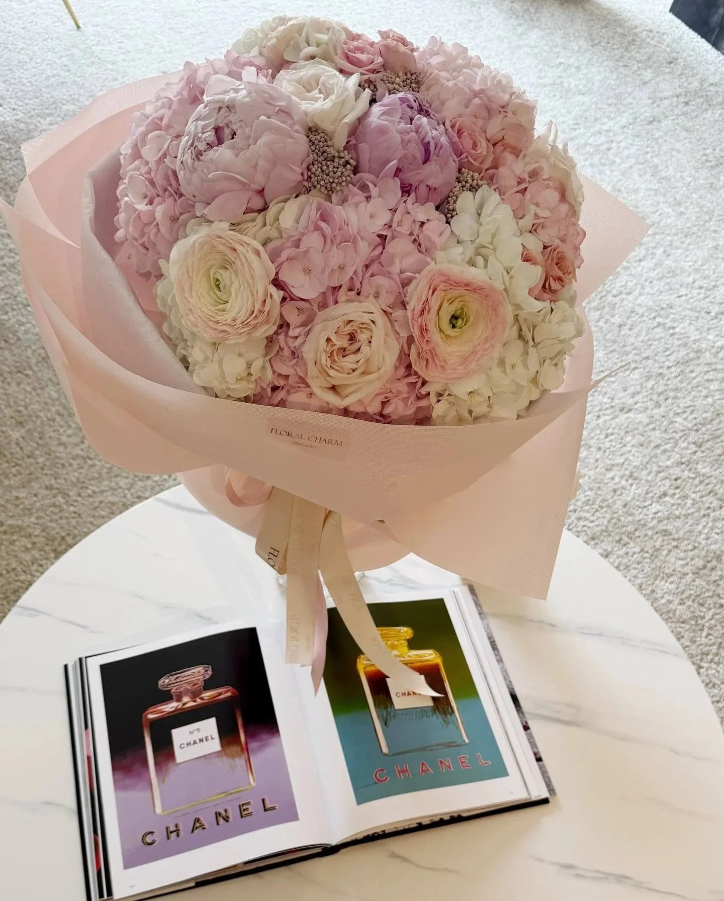 A bouquet of pink and white flowers, including roses and hydrangeas, wrapped in pink paper, sitting on a white table next to an open Chanel perfume magazine.