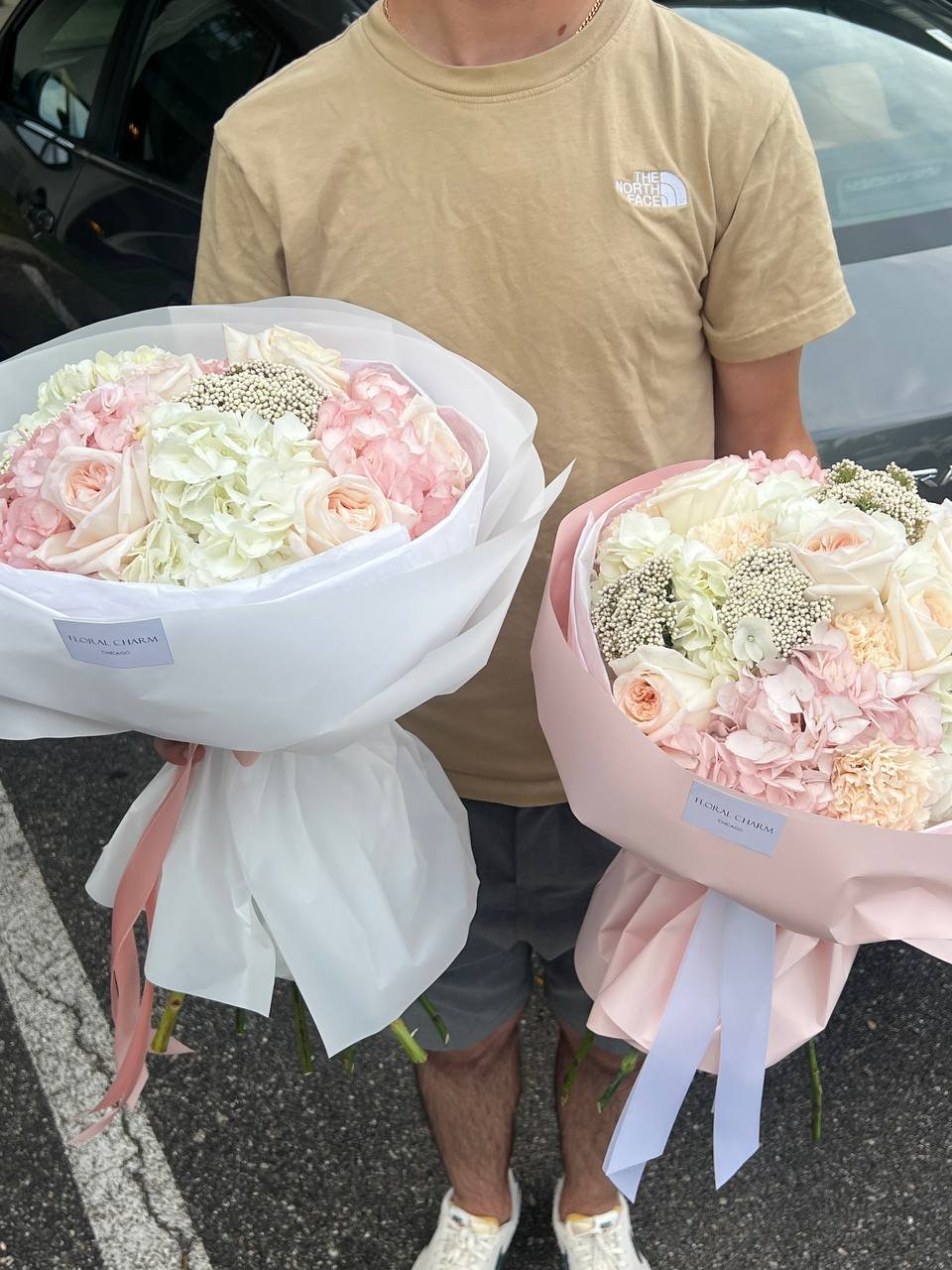 Person holding two large bouquets of pink, white, and peach flowers in a parking lot.